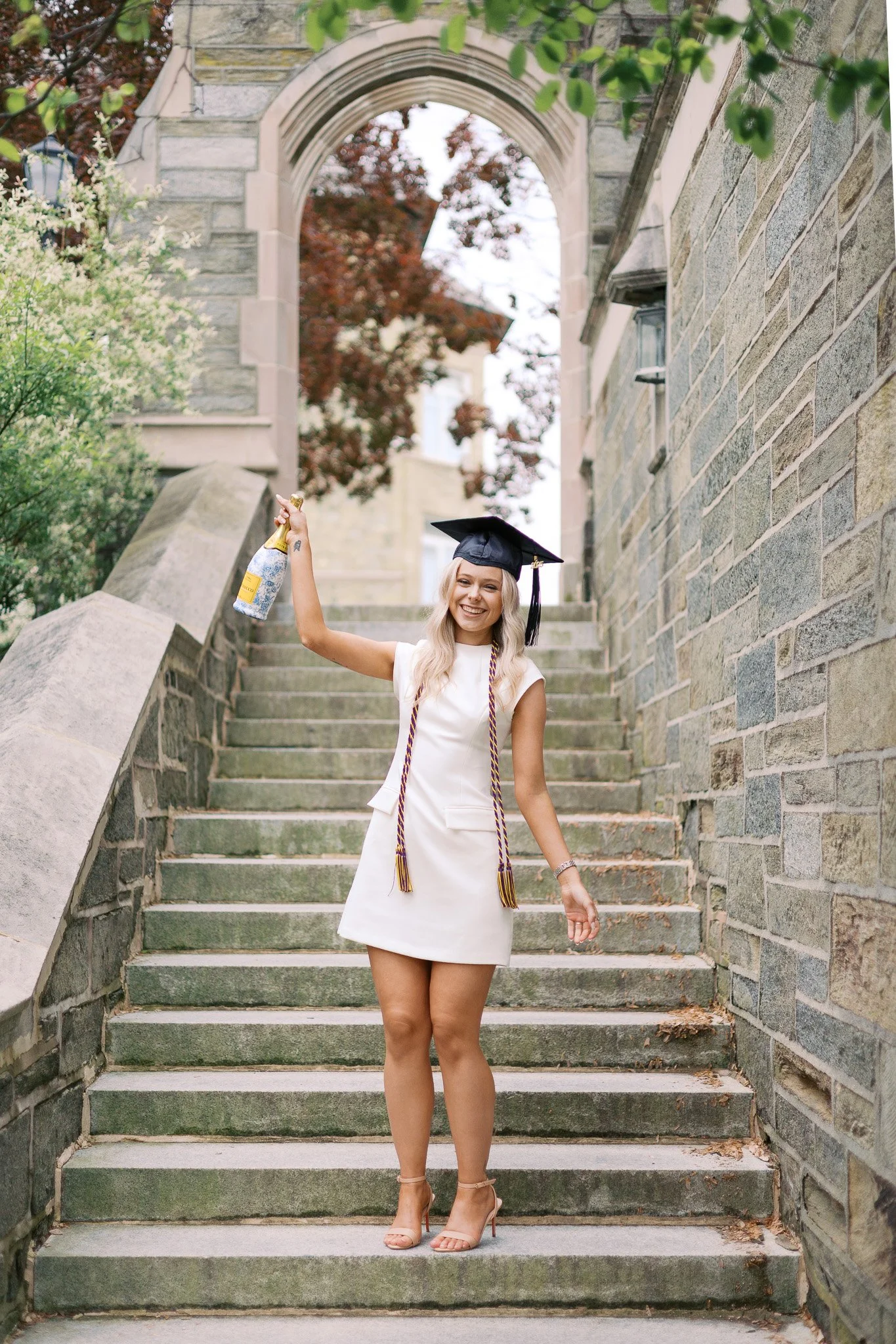 A young WCU student with blonde hair, wearing a white dress, high heels, and a graduation cap, standing on stone stairs outside, celebrating with a bottle of champagne and smiling.