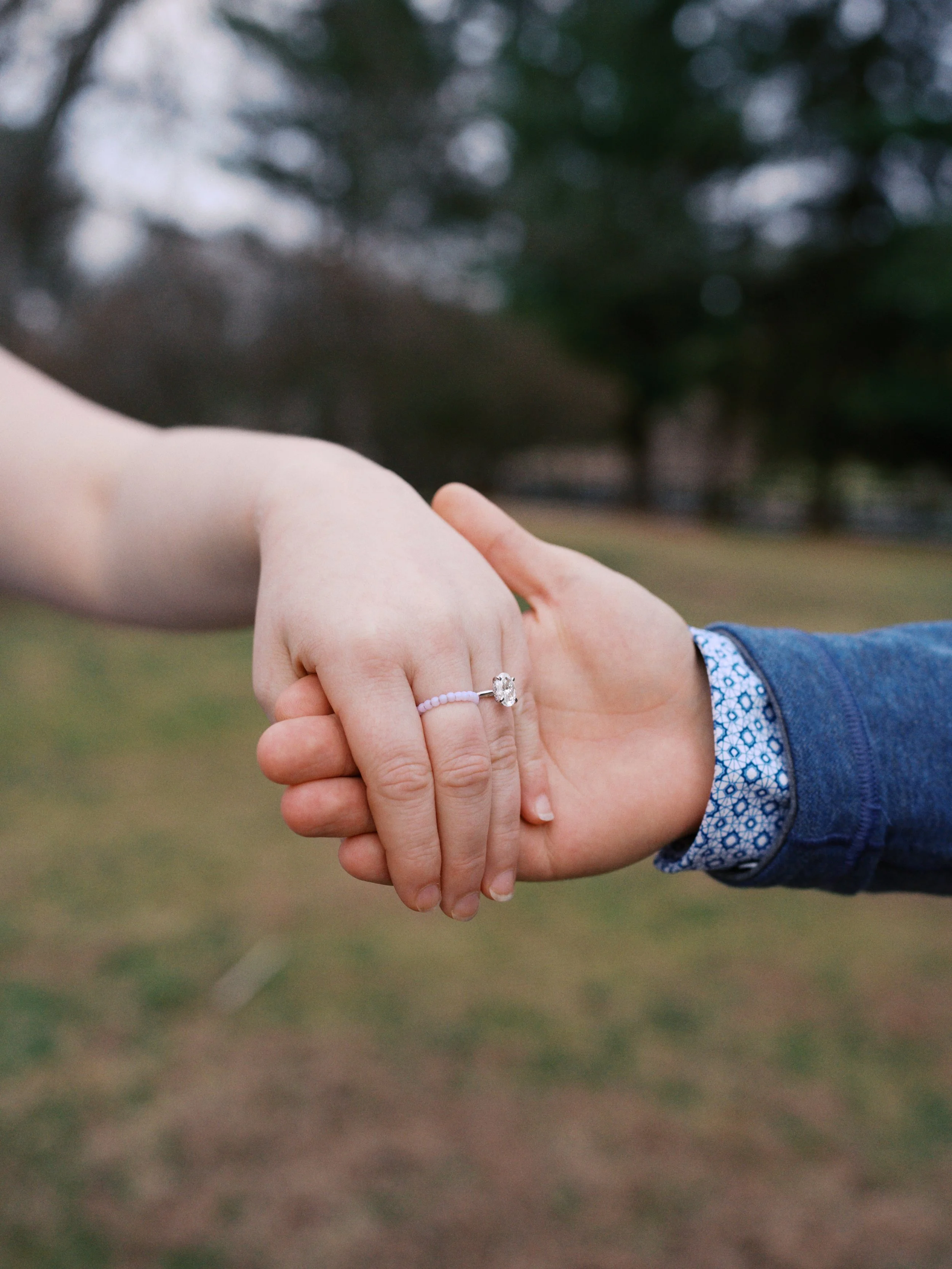 Close-up of a couple holding hands outdoors, with focus on a woman's hand wearing an engagement ring, and a man's hand gently holding hers.