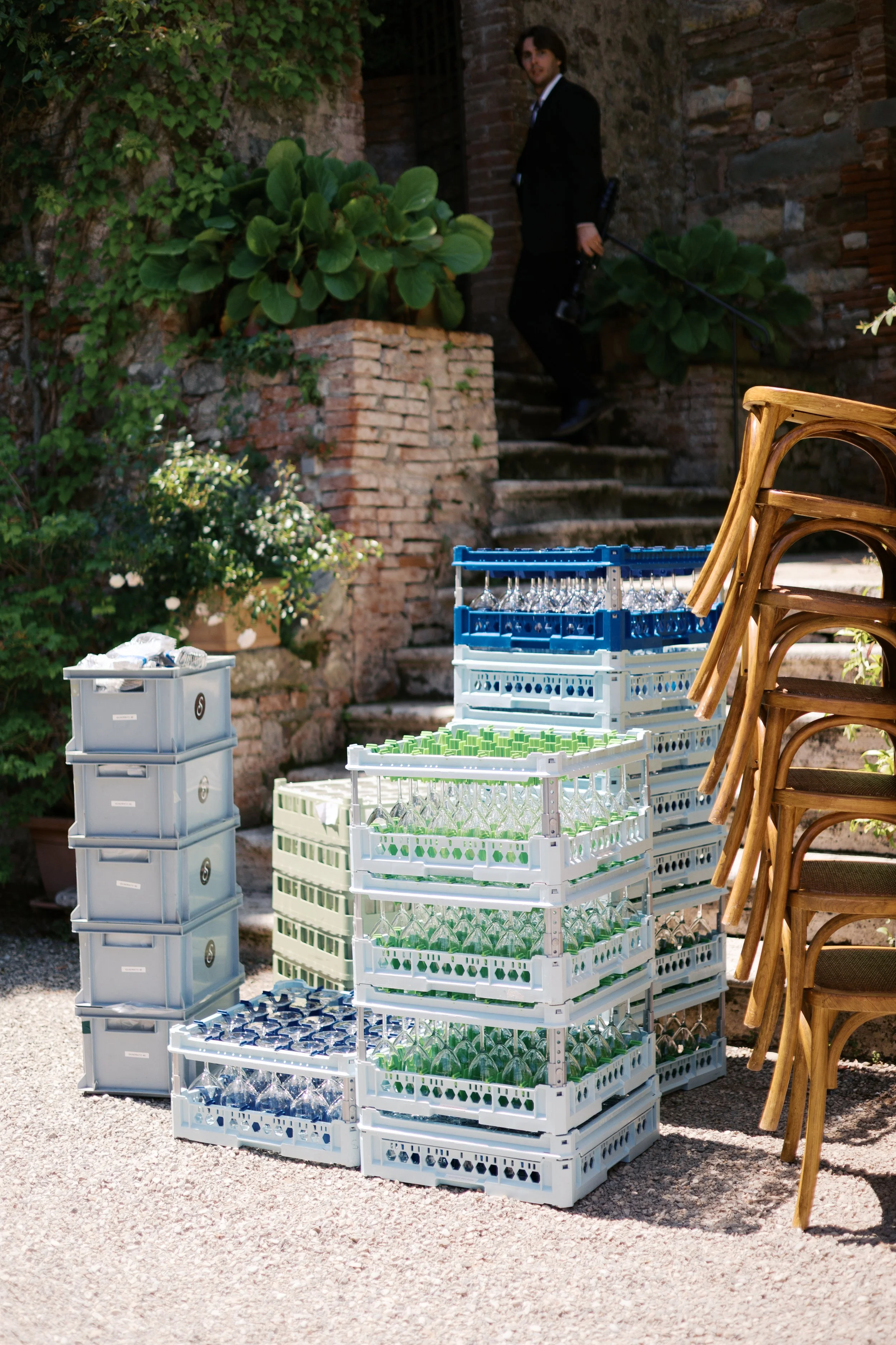 Plastic crates filled with glass bottles are stacked outdoors on a stone ground near some wooden chairs. A man in a black suit is descending stone stairs, holding a camera, in the background.