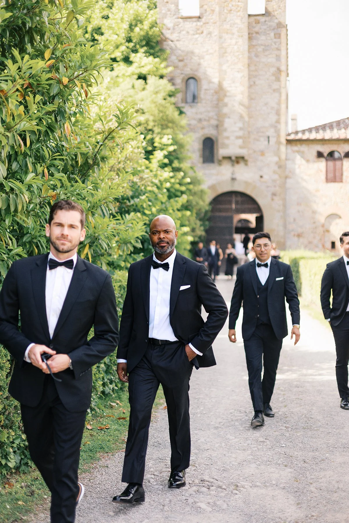 Group of men in tuxedos walking outdoors near a stone building and green bushes.