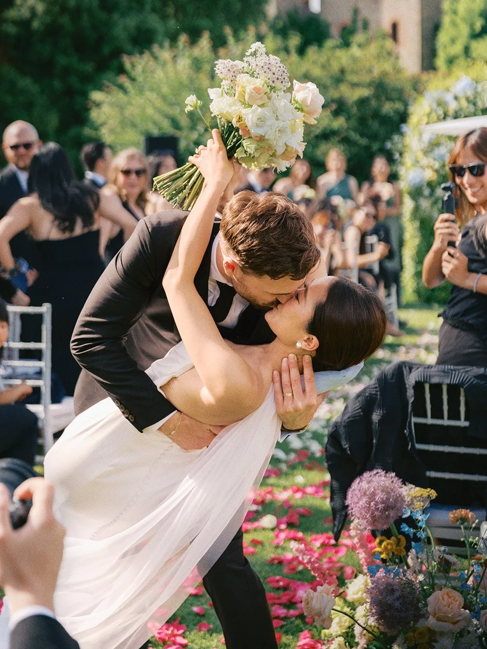 A bride and groom in Tuscany, Italy sharing a kiss during their wedding ceremony outdoors, with the groom holding a bouquet over the bride's shoulder, surrounded by guests and flower petals on the ground.