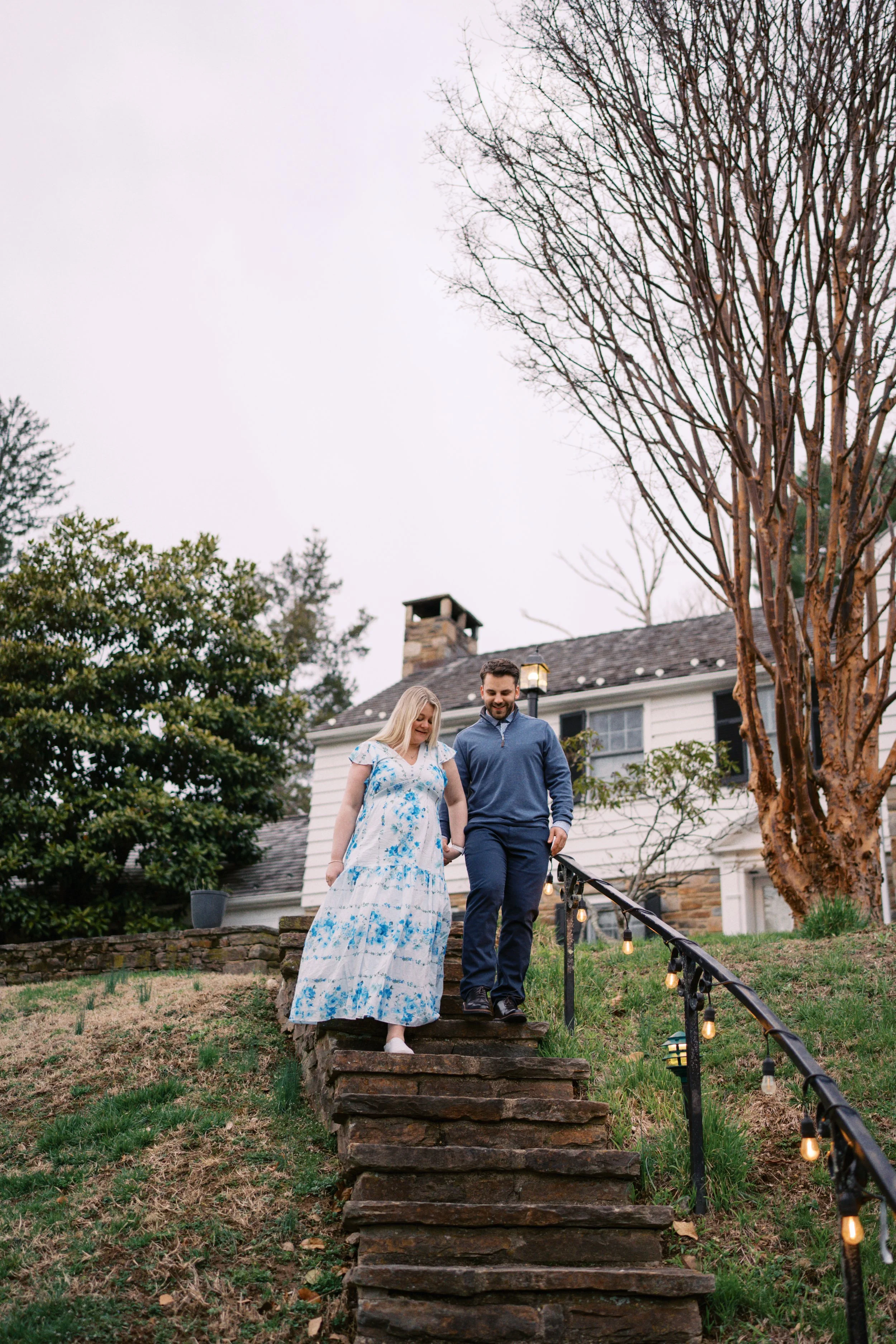 A man and woman, holding hands, walk down stone steps outside a white house with a chimney. The woman wears a white dress with blue floral patterns, and the man wears a blue jacket and dark pants. There are string lights on the railing and trees in the yard.