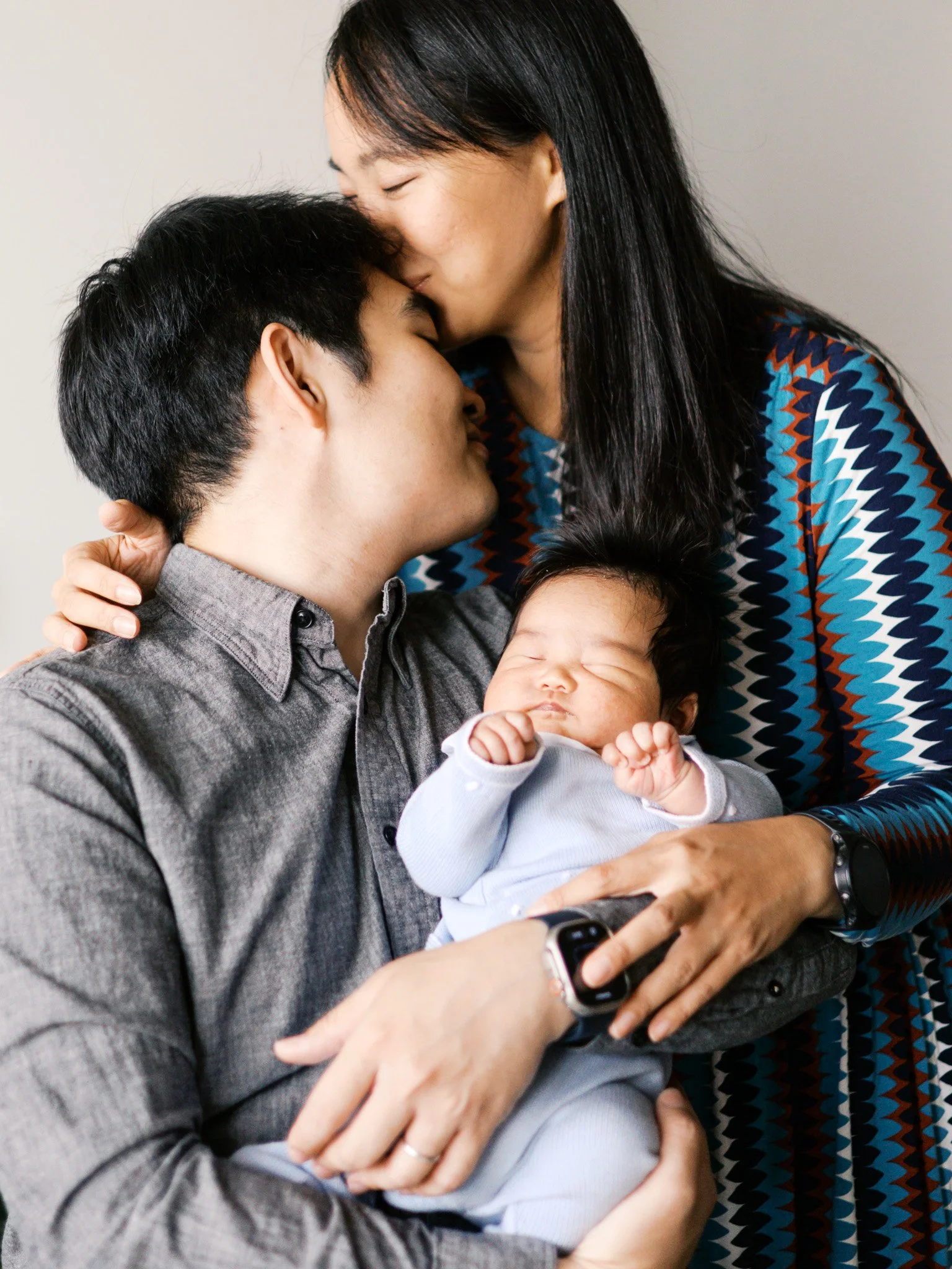 A family of three, with a woman kissing a man on the forehead while he holds a sleeping baby in his arms.