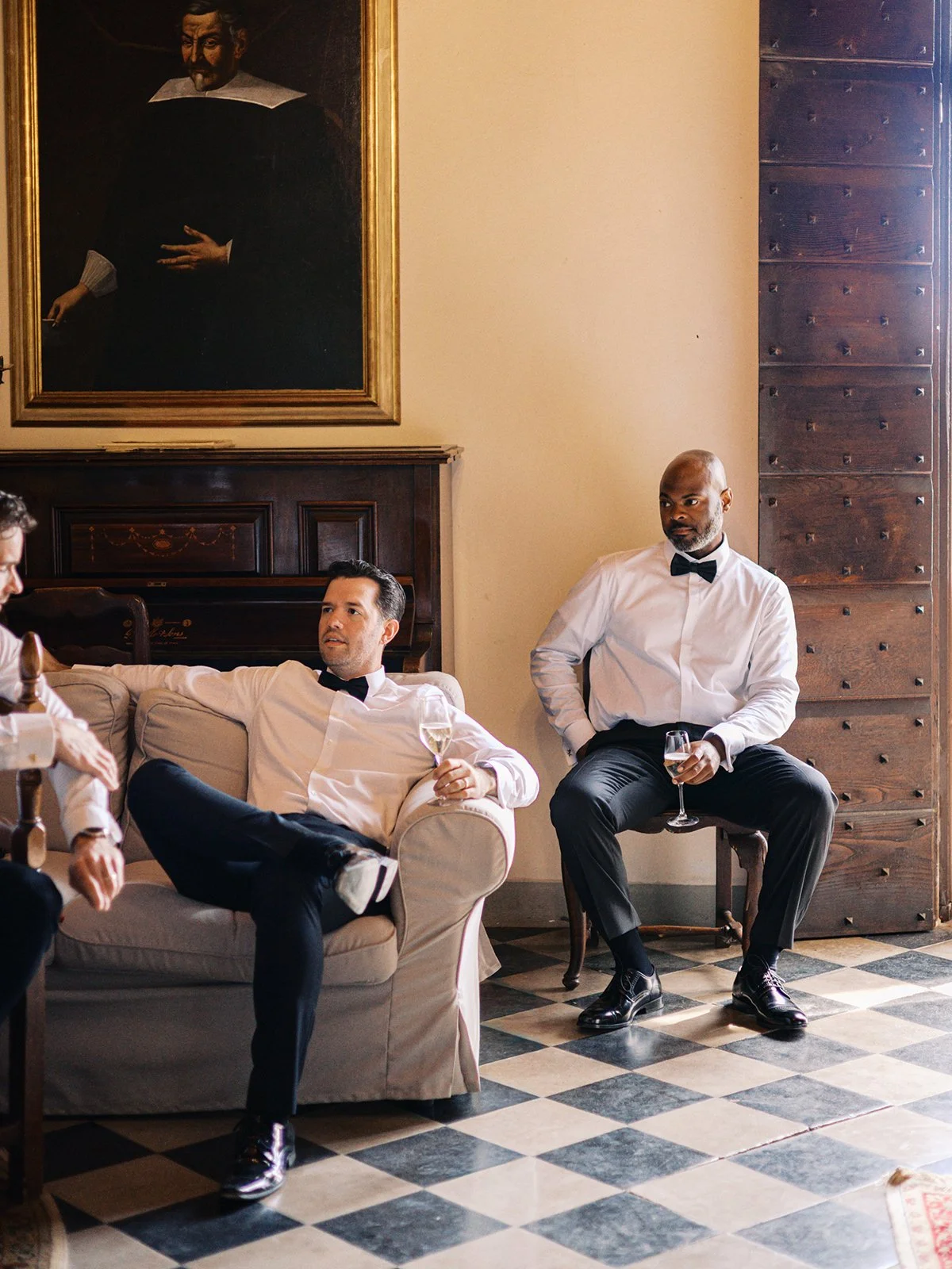 Group of men dressed in formal attire, sitting and standing in a vintage-style room with a portrait hanging on the wall.
