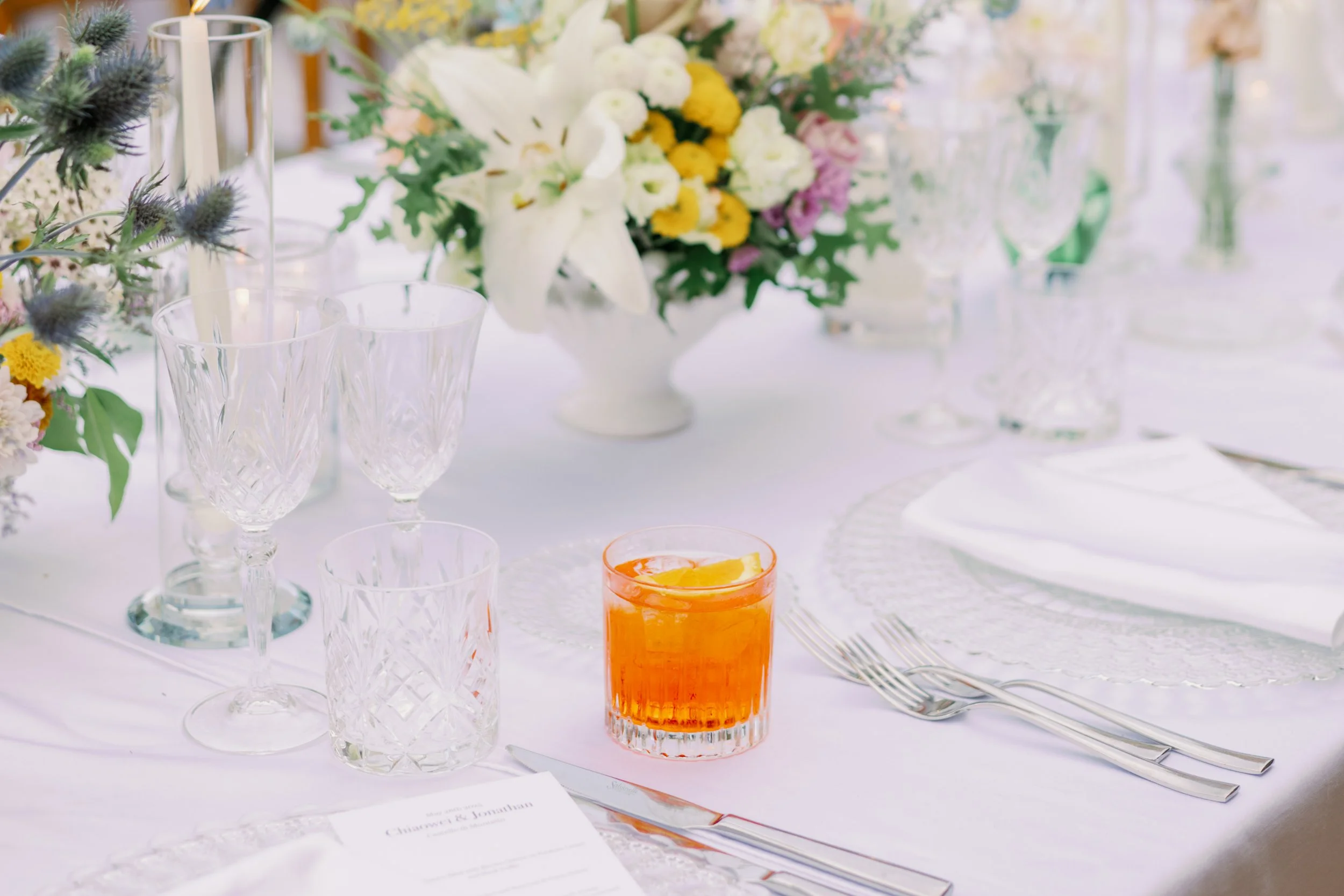Elegant table setting with crystal glasses, a floral centerpiece with white lilies, yellow and purple flowers, a drink with a lemon slice, and neatly arranged cutlery.