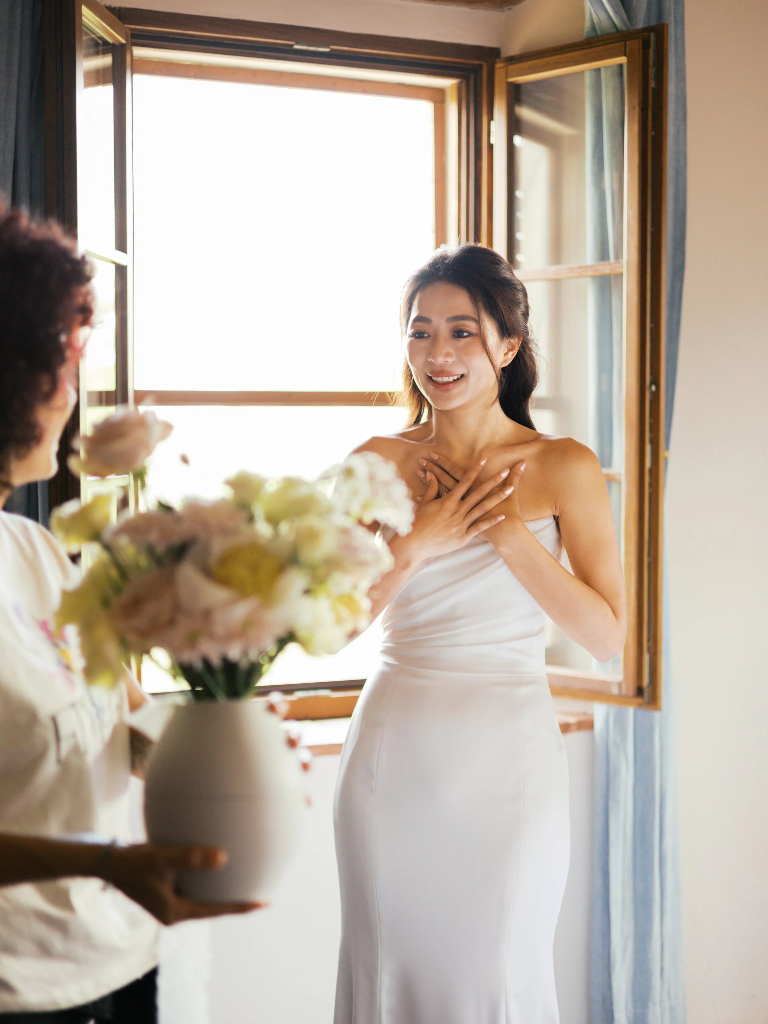A bride in a white dress with her hands on her chest, smiling, standing near an open window, with another person holding a bouquet of flowers nearby in Tuscany, Italy.