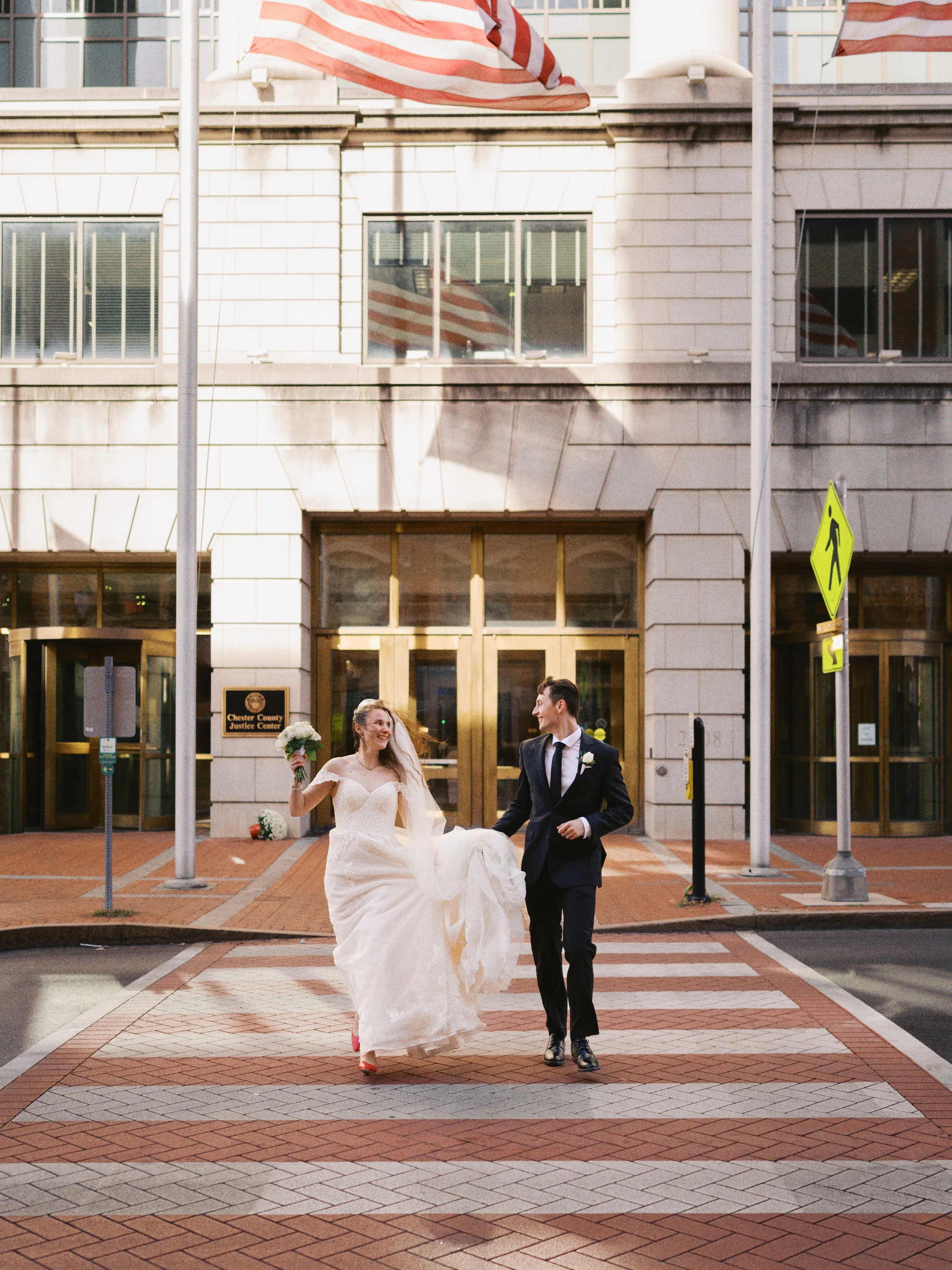 Bride and groom walking across a crosswalk in front of the Chester County Justice Center. The bride is wearing a wedding gown and holding a bouquet, while the groom is in a suit. They appear happy and are smiling at each other.