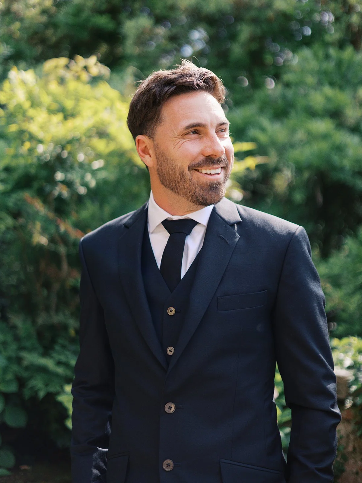 A man in a dark suit and tie standing outdoors amidst greenery, smiling happily.
