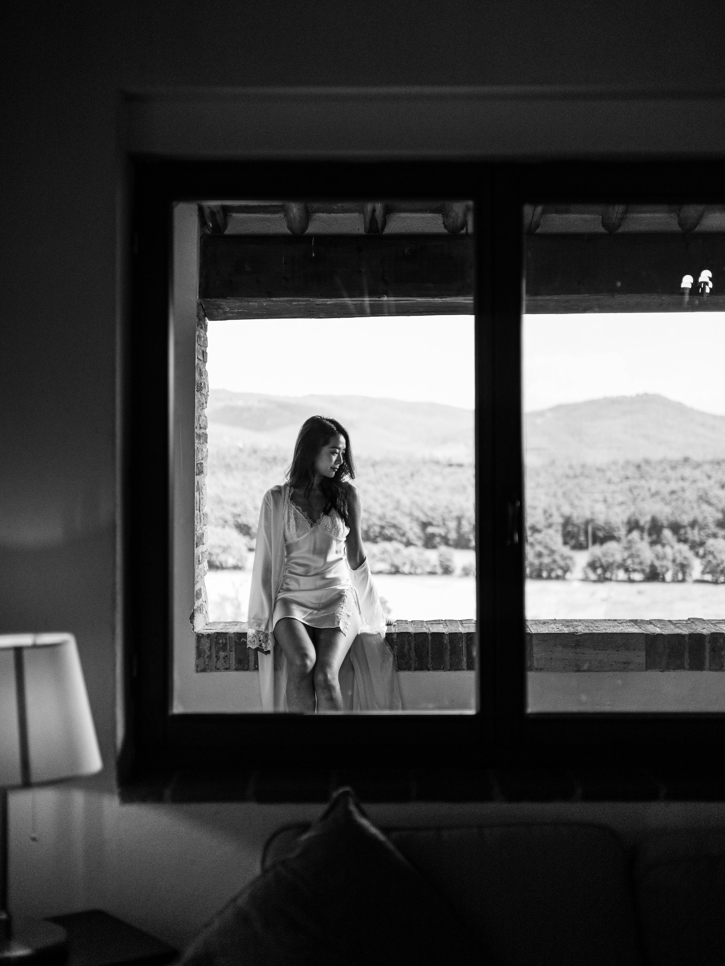 A woman sitting on a window ledge, wearing a silky dress, looking down with mountains and trees in the background.