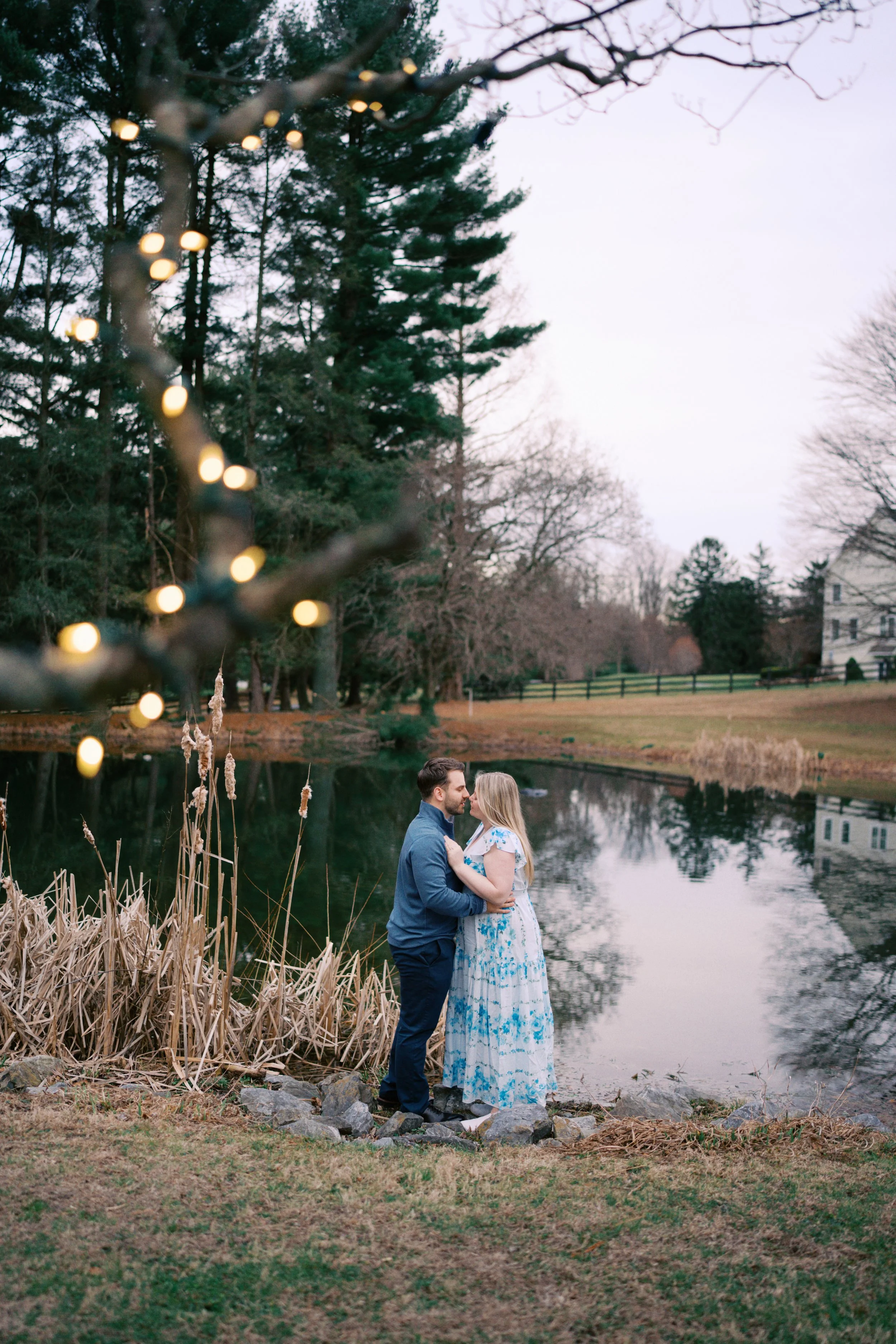 A man and woman standing close together by a pond in West Chester, PA, holding hands, with trees and a house in the background, during dusk.