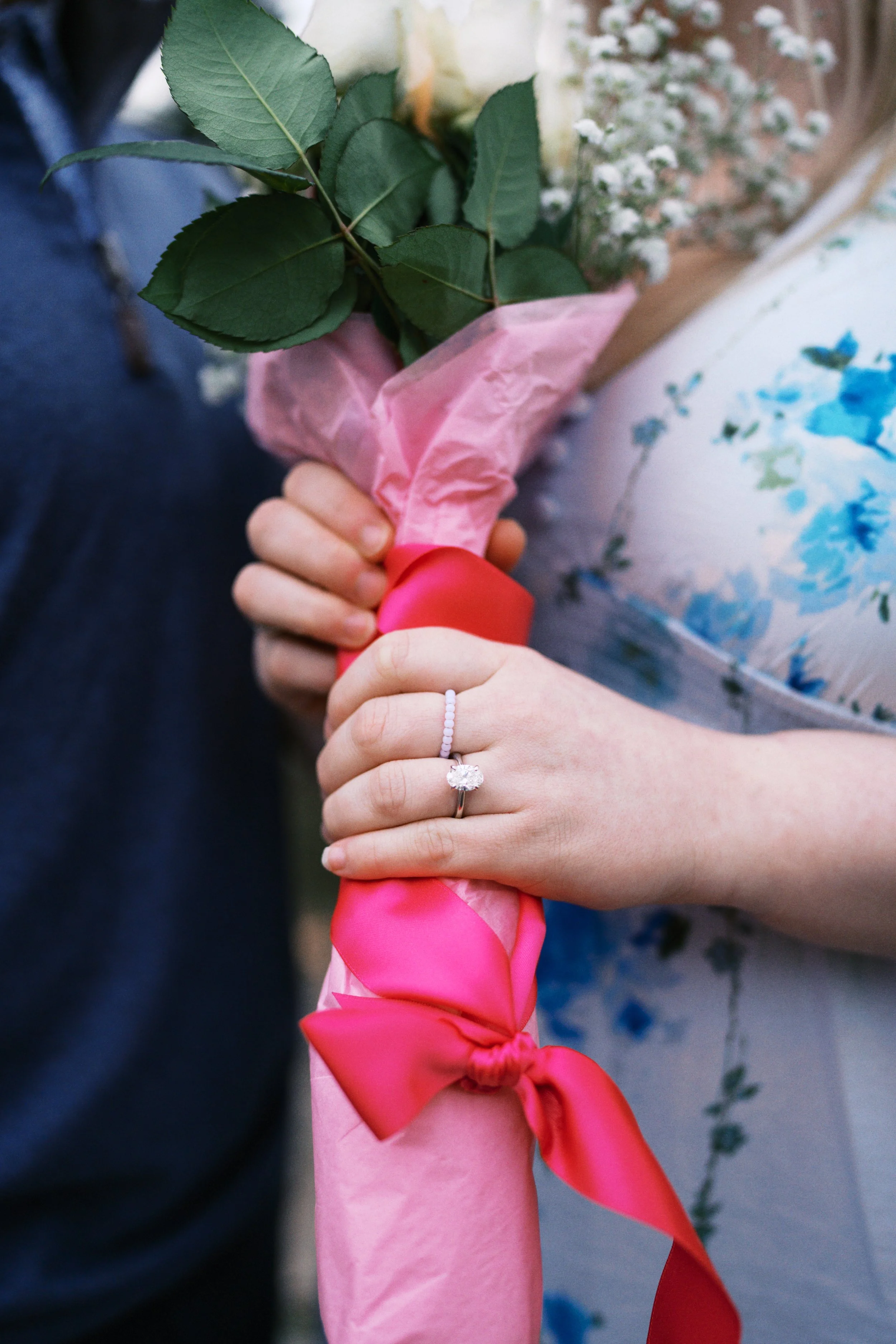A person holding a bouquet of white flowers wrapped in pink tissue paper, with a ribbon. The person is showing an engagement ring on their finger.