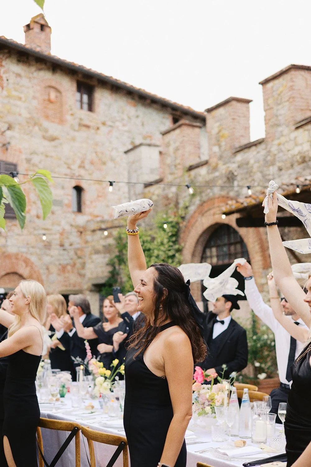 People celebrating at a wedding reception, raising napkins and clapping, seated at a long table with flowers and drinks, outdoors with stone buildings and string lights in the background.