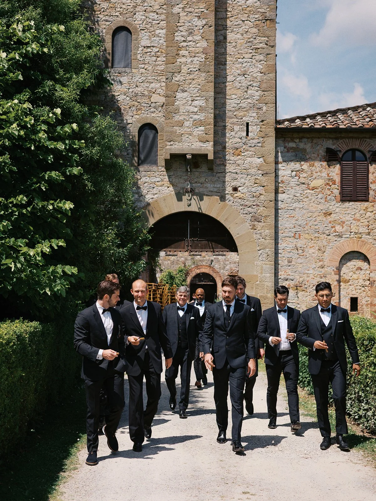 A group of men in formal suits, walking outdoors near a stone building with an archway, on a sunny day.