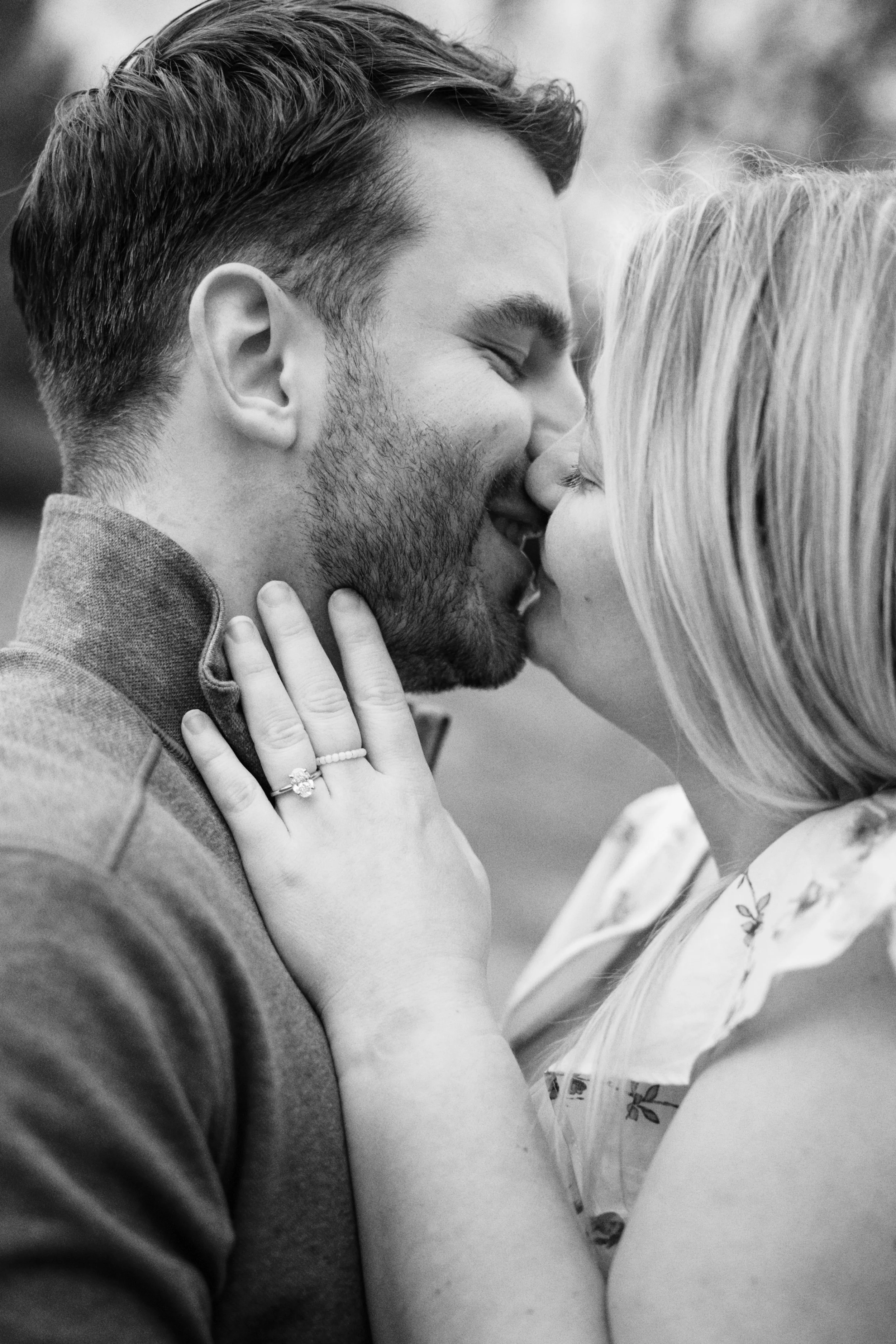 A black-and-white photo of a couple sharing a kiss, with the woman holding the man's face and showing an engagement ring on her finger.