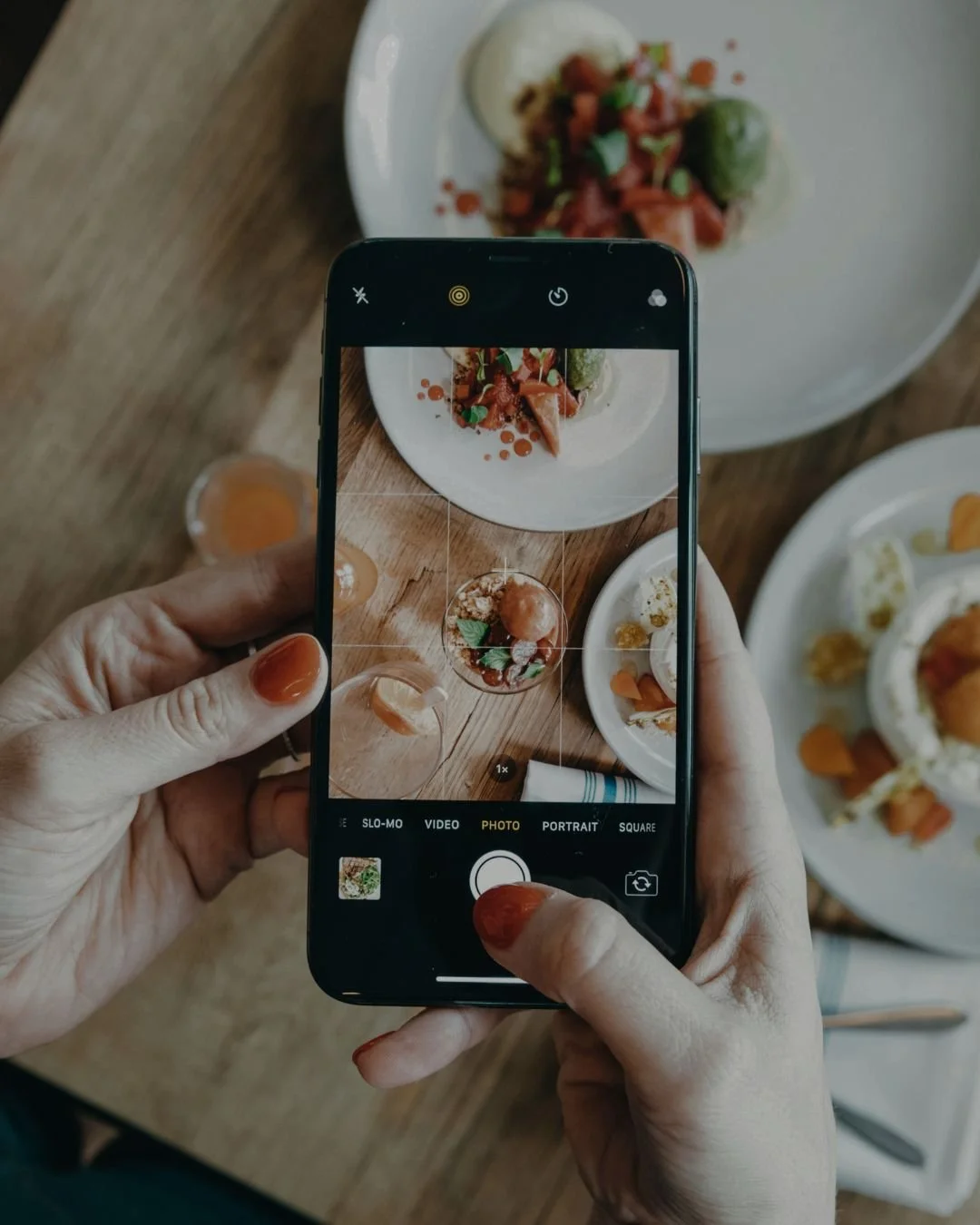 Person taking a photo of a gourmet dish with a smartphone. The dish features diced tomatoes, herbs, and a dollop of white sauce or cream, served on a white plate.