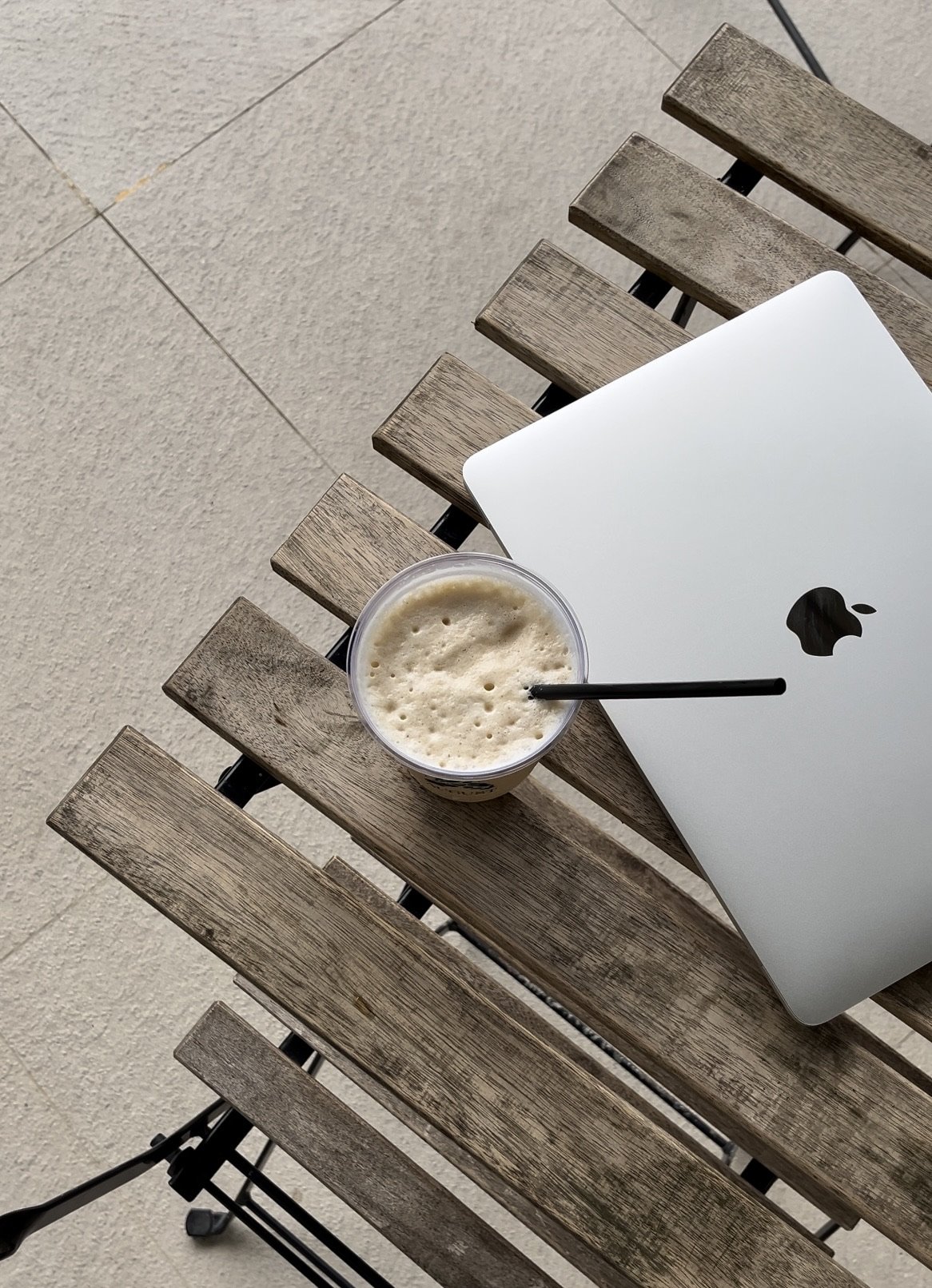 A laptop, a cup of frothy beverage with a straw, placed on a wooden outdoor table.
