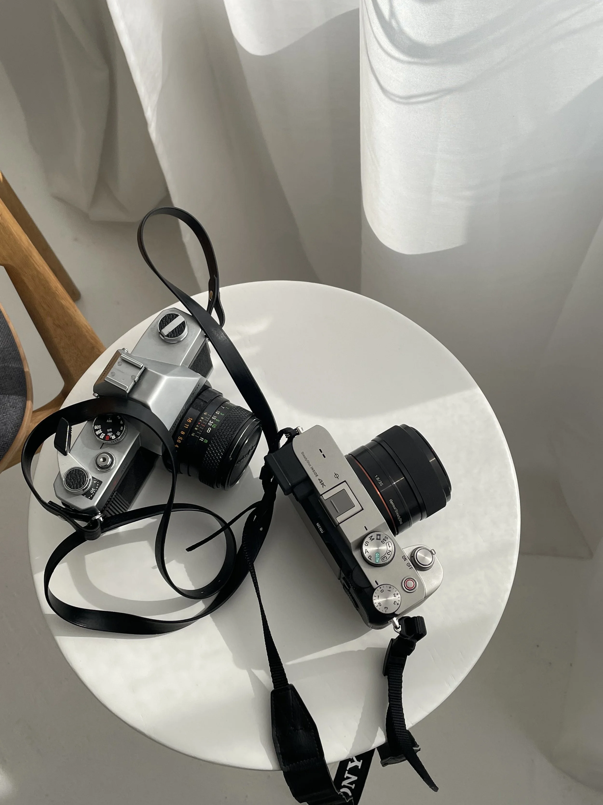Two cameras, one silver and black DSLR and one silver compact, resting on a white round table with a white curtain in the background.