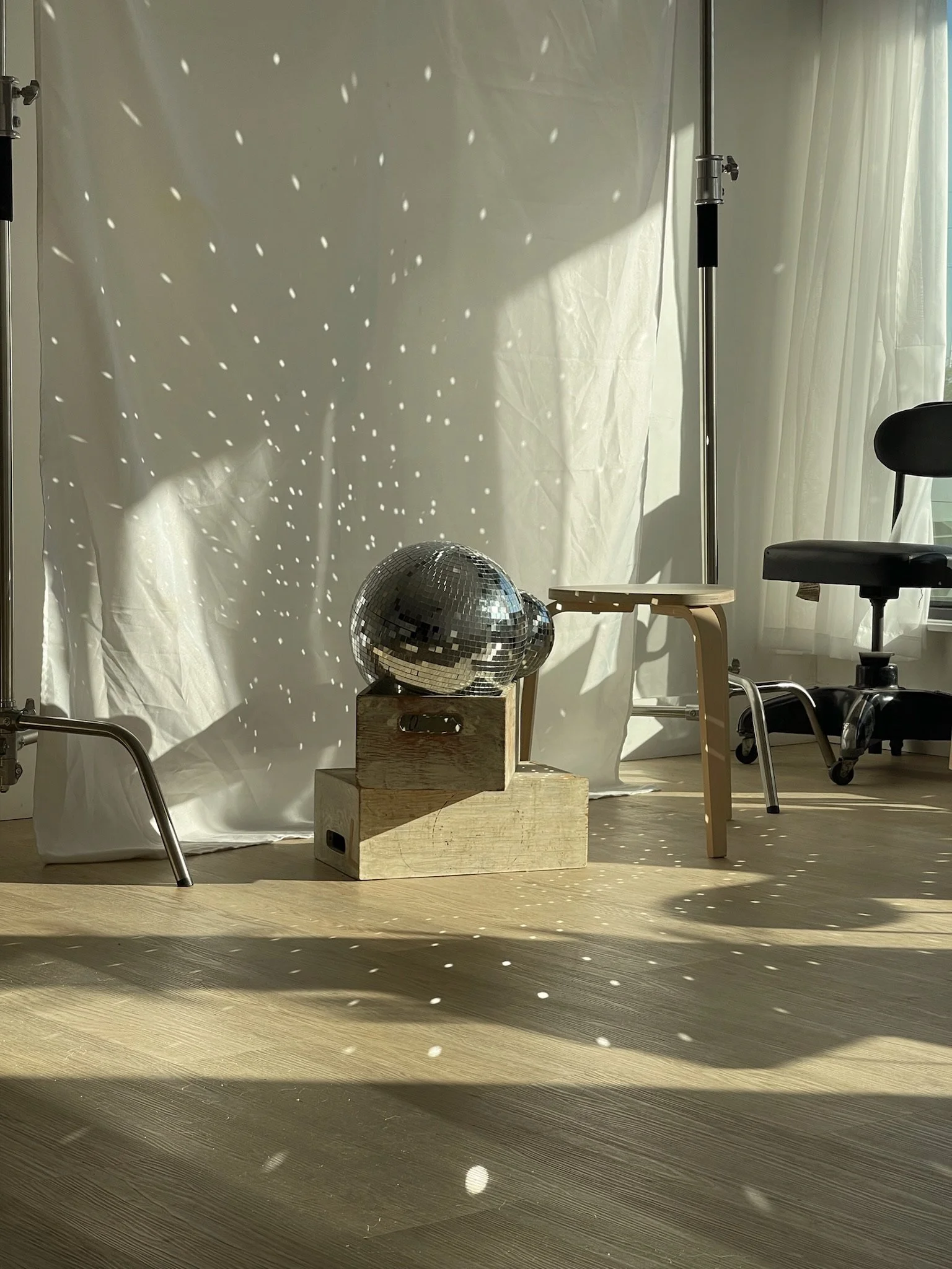 Room with sunlight creating reflections on a white backdrop, featuring a disco ball on a wooden box, a small beige bench, and a black office chair.