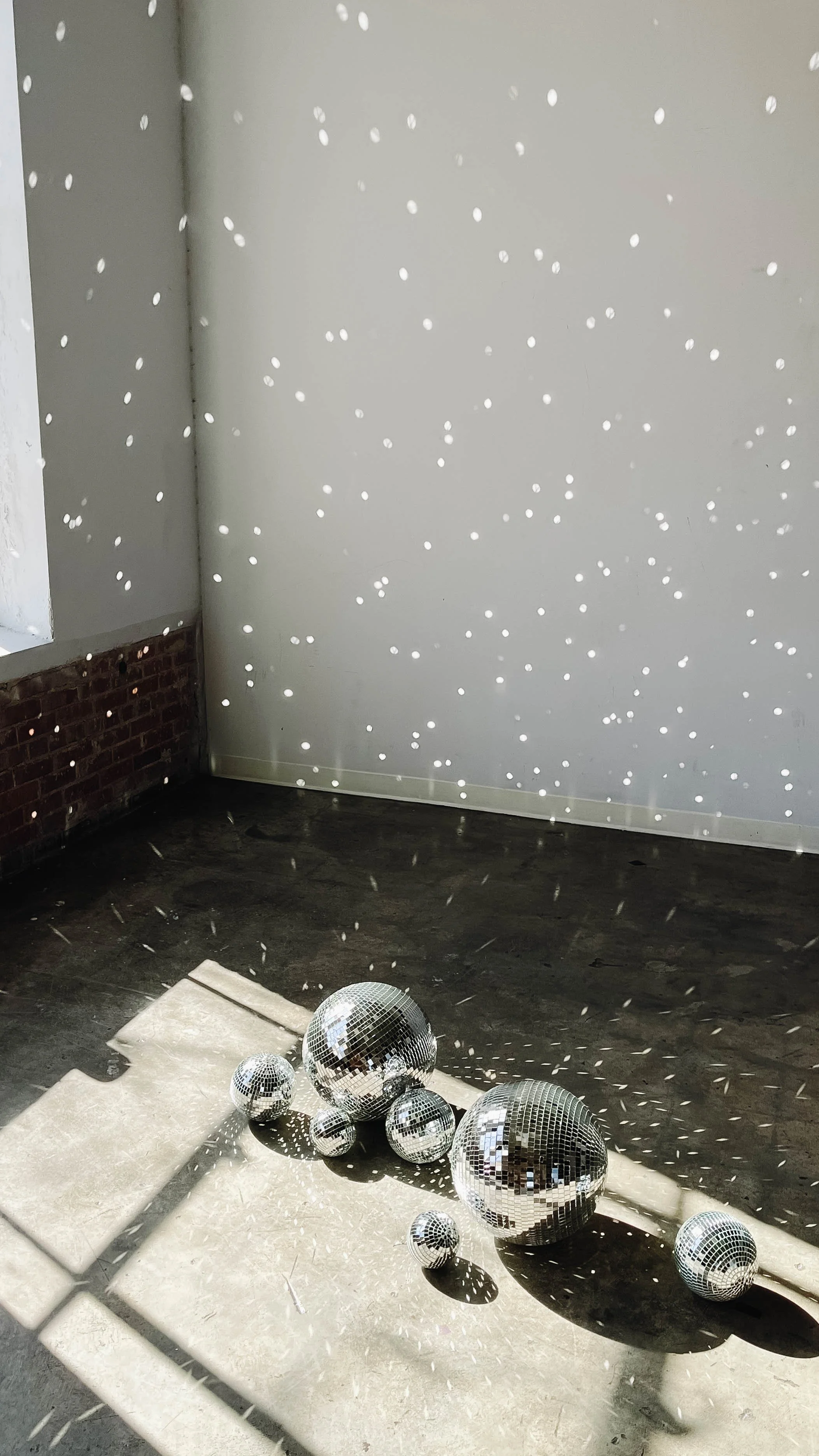 Disco balls reflecting light in a room with concrete floor and white walls, creating a dotted light pattern on the walls and floor.