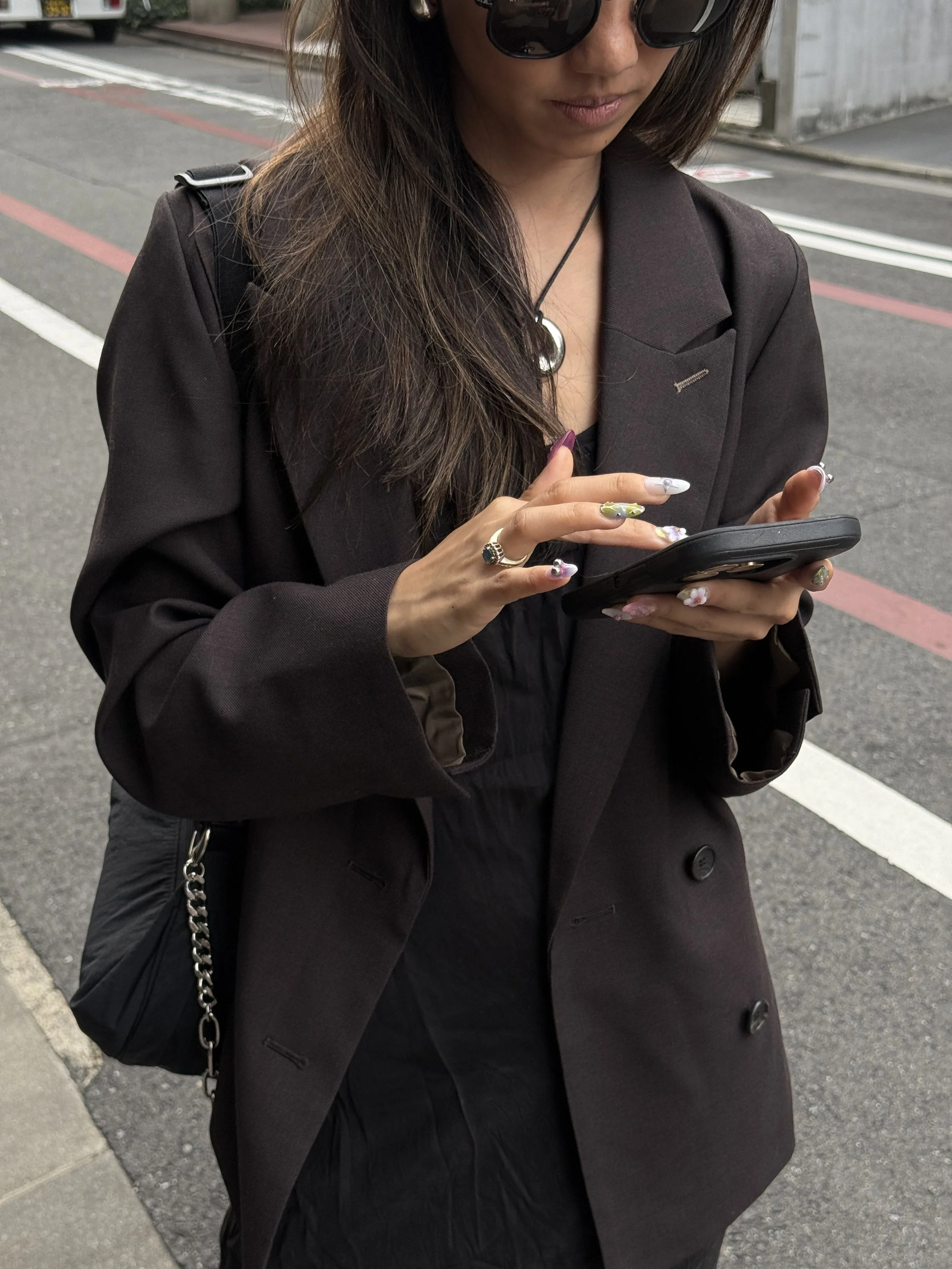 A woman standing outdoors on a city street, wearing a dark brown blazer and black dress, with long hair, sunglasses, and jewelry, looking at her phone.