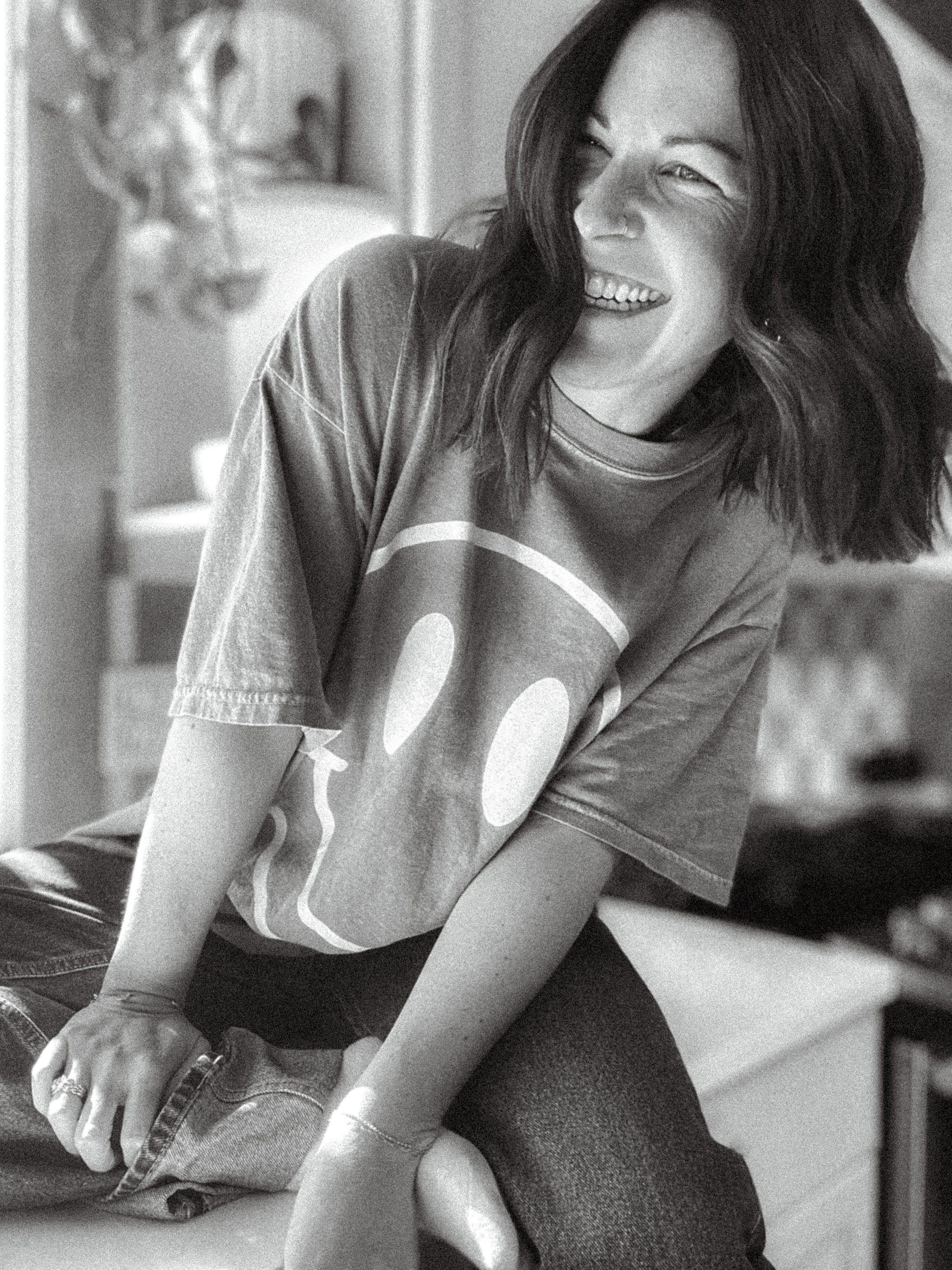 A woman with dark hair smiling and laughing, wearing a graphic T-shirt, sitting on a bed in a bedroom.