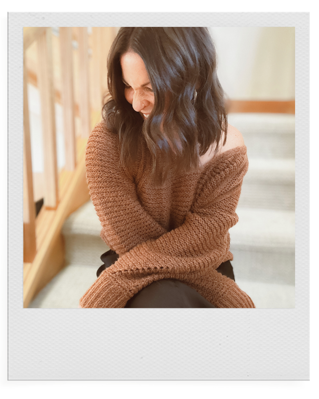 A woman with dark, wavy hair laughing and leaning forward, wearing an oversized brown sweater, sitting on stairs with wooden railing in background.