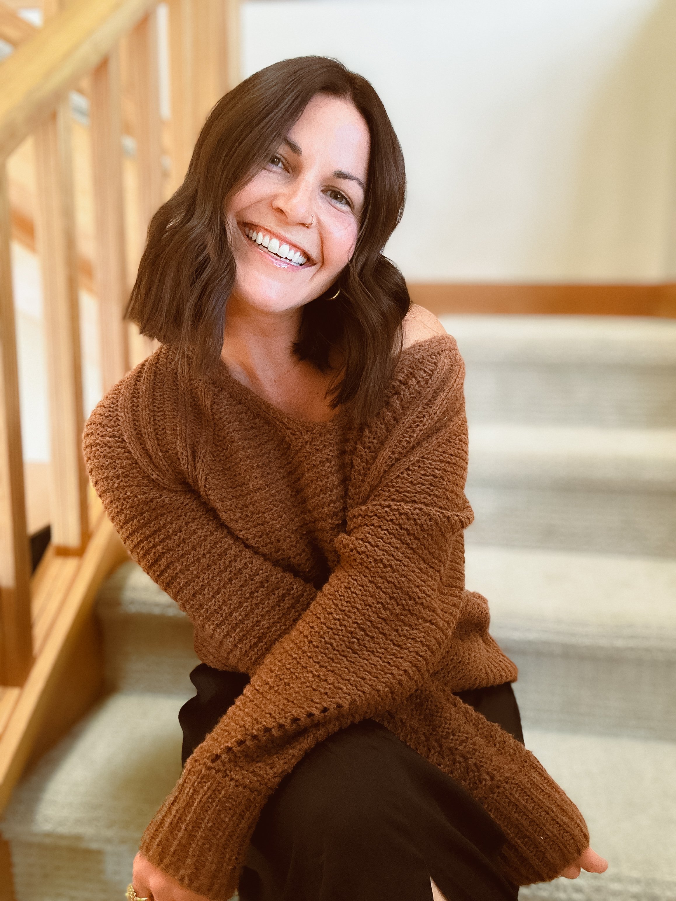 A woman with shoulder-length brown hair, wearing a brown knitted sweater, sitting on stairs and smiling at the camera.