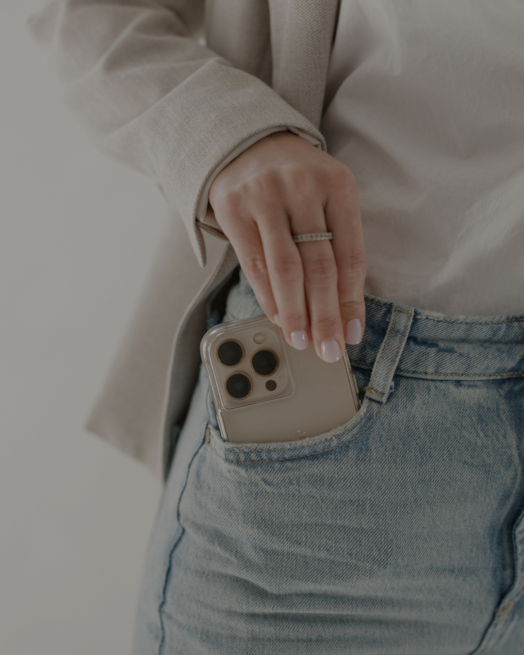 Close-up of a person with a ring on their finger, wearing a beige blazer and light blue jeans, placing a smartphone into their front pocket.