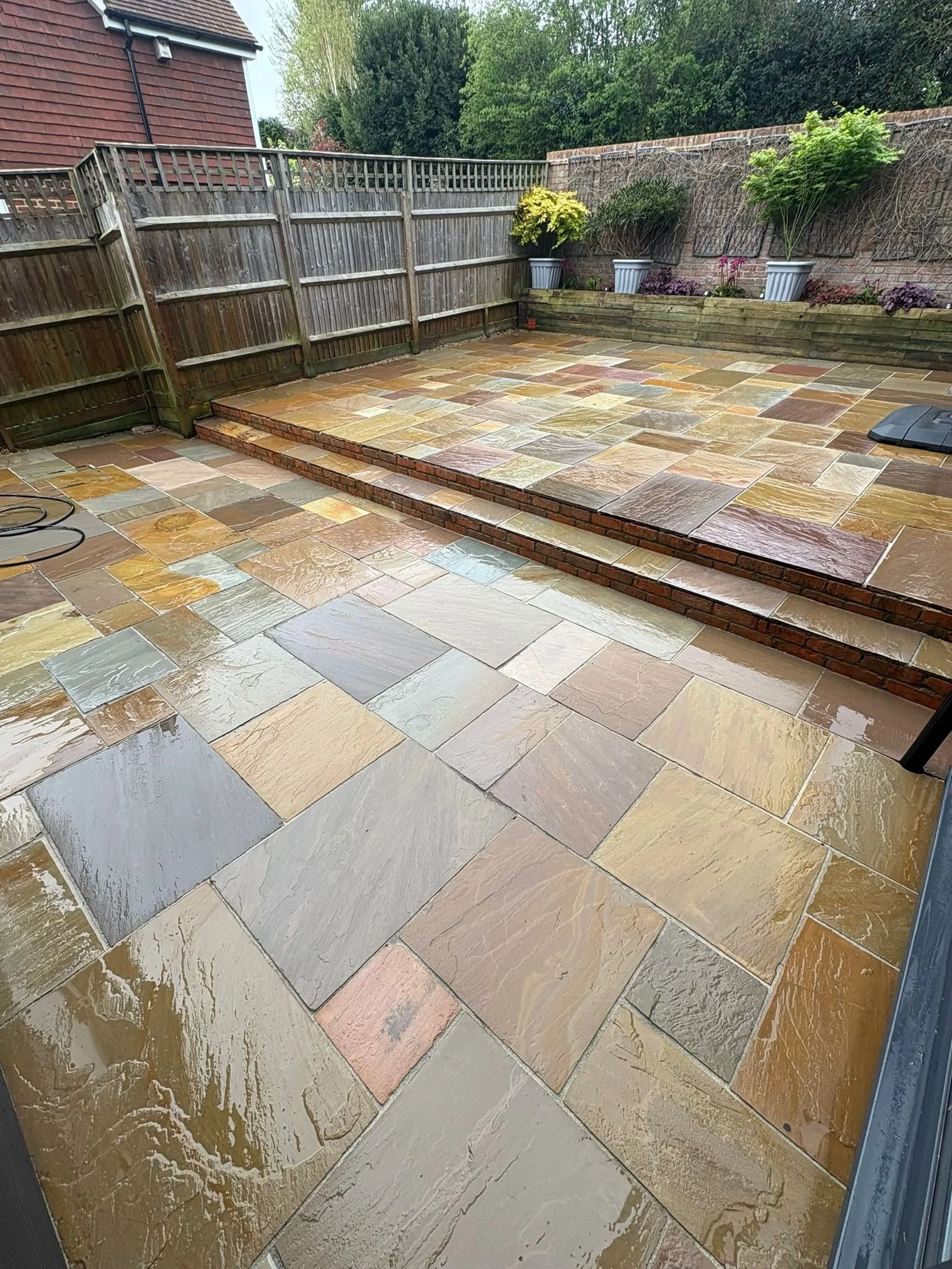 A wet, multicolored stone patio with steps leading up to a higher level. A wooden fence runs along the back, with potted plants and greenery behind it.