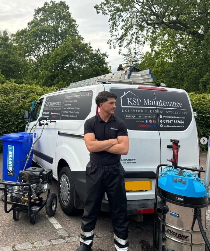 A man in black uniform standing in front of a white van with a company logo for KSP Maintenance, exterior cleaning specialists. There are cleaning equipment, a blue water tank, and a machine on a cart nearby. The background features trees and a cloudy sky.