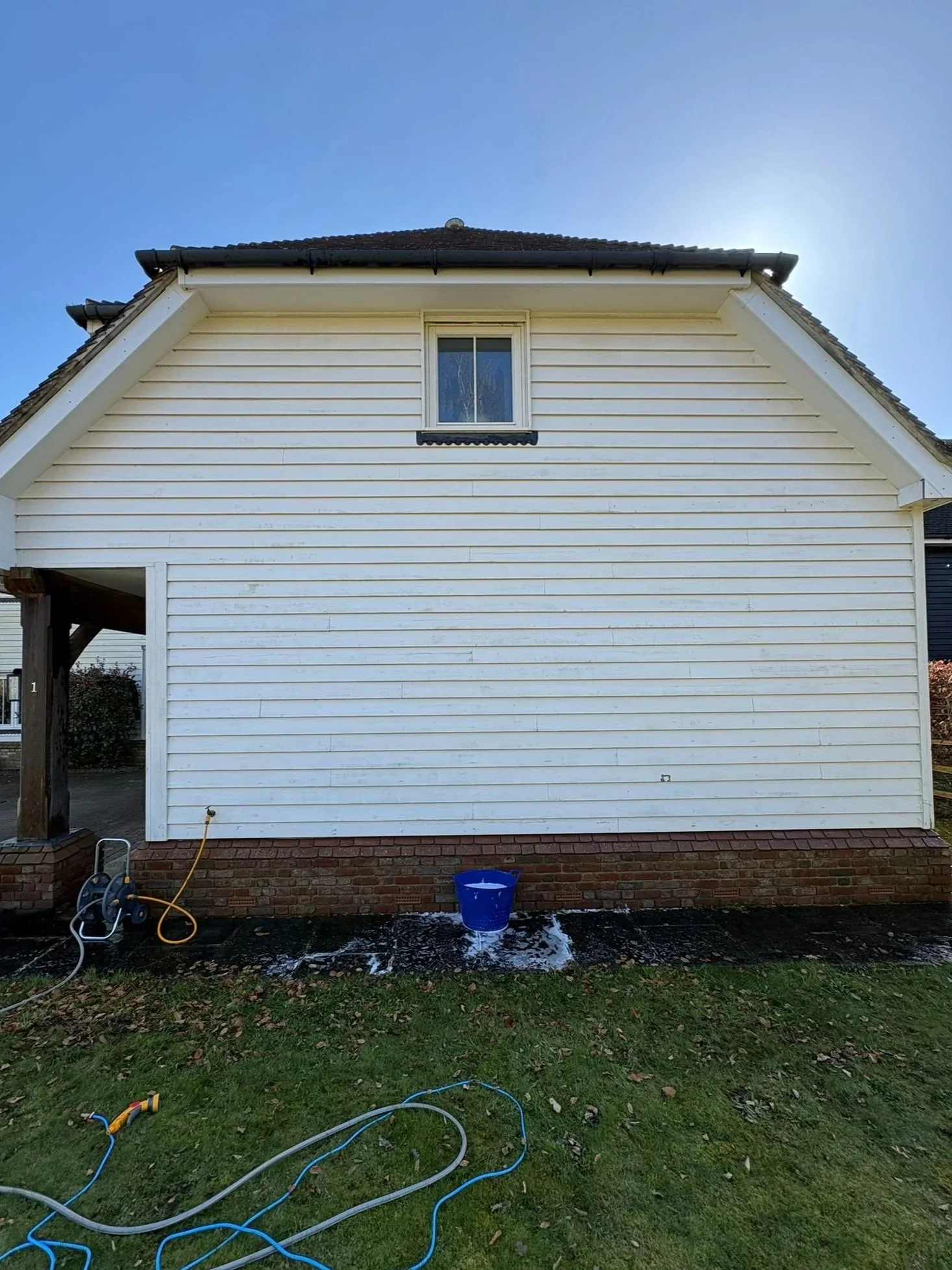 Back of a house with white wooden siding, a small window, and a brick foundation. There is a blue bucket on the ground with water underneath, and garden hoses and cleaning supplies nearby, suggesting house cleaning or maintenance activities.