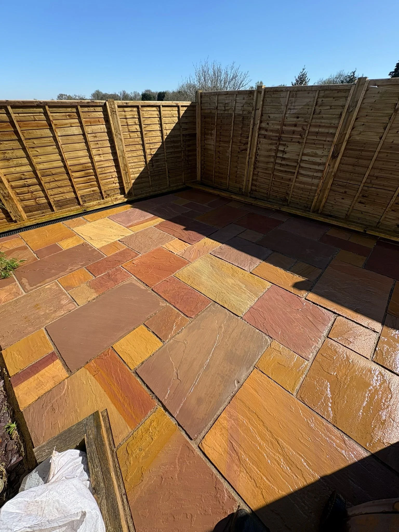 A wooden fence with a diagonal shadow, a tiled patio with multicolored stone tiles, and a blue sky with some trees in the background.