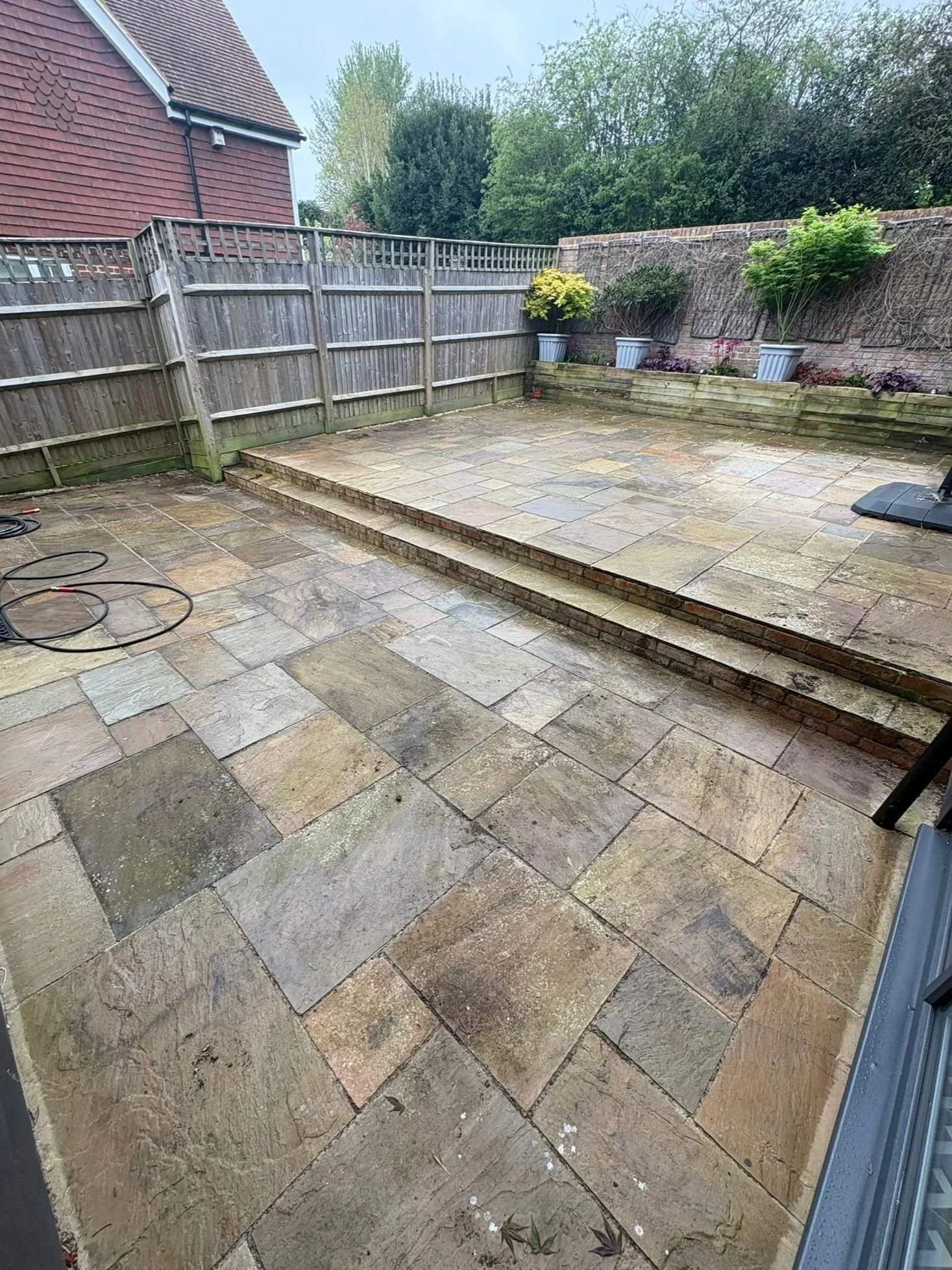 Clean outdoor patio with large stone tiles, brick steps, and potted plants against a growth of trees and a brick wall.