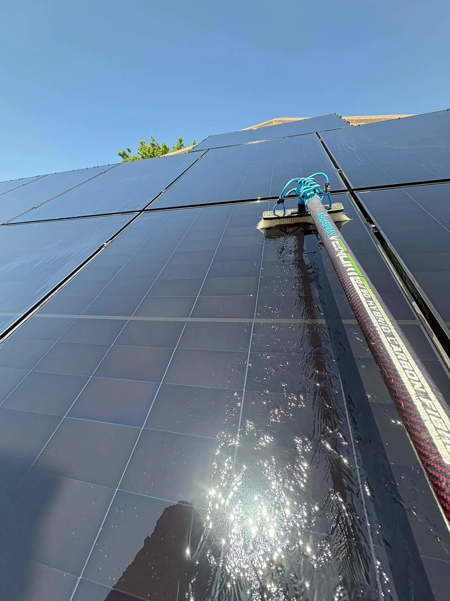 A person cleaning or maintaining solar panels on a building roof, with a clear blue sky and some greenery visible in the background.