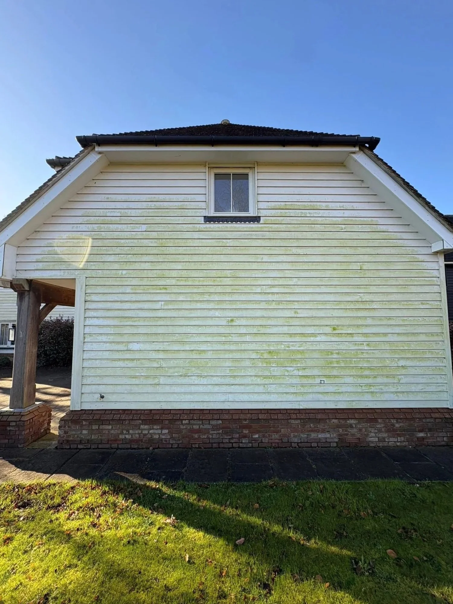 Side view of a house with white siding, a small window, a brick foundation, and some green moss or algae stains near the top. The house has a black roof and part of a wooden post and porch is visible on the left. There is a grassy area with sunlight below the house.
