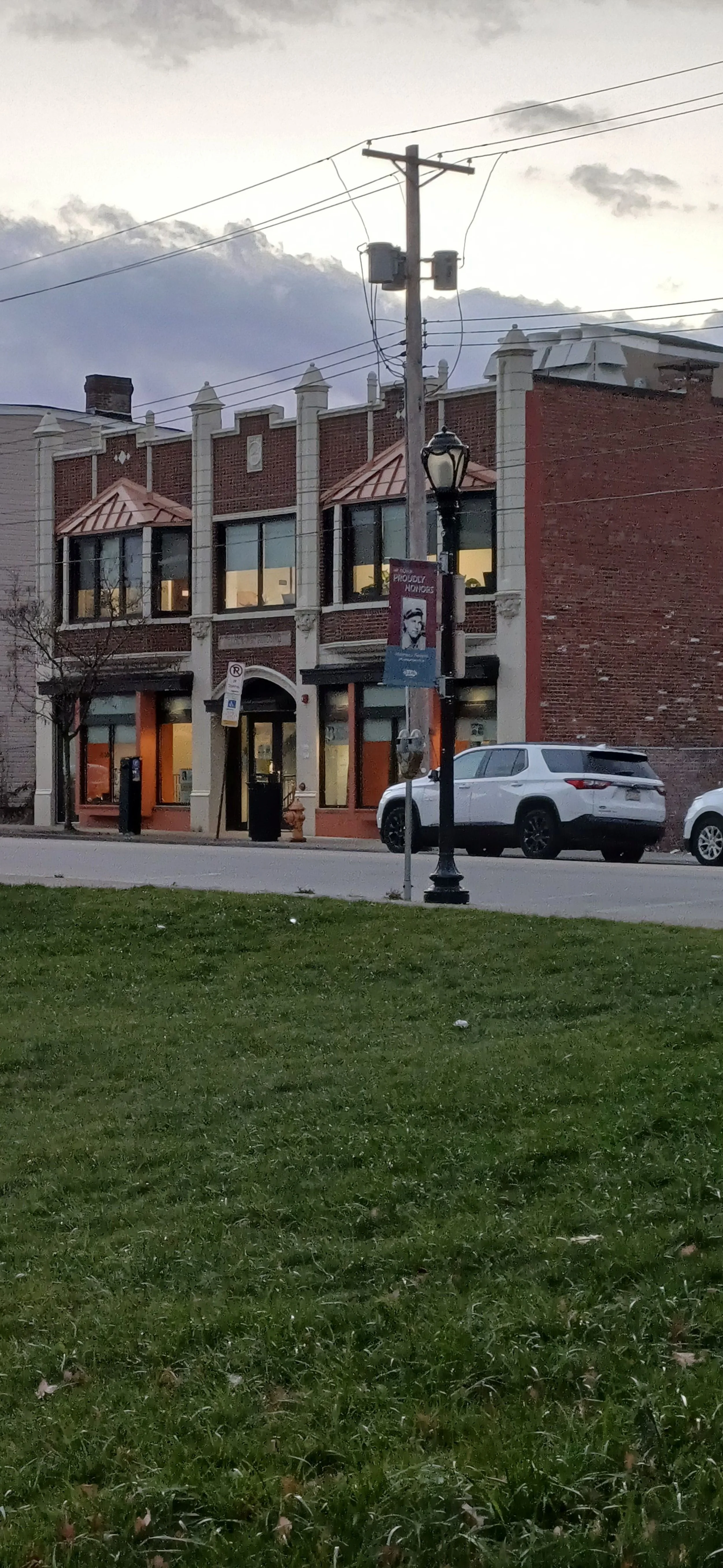MT Oliver Pa street scene with a brick building featuring large windows on the second floor, a small storefront on the ground floor, a parking meter, a sign, and parked cars, with power lines overhead and a lamppost, during dusk or early evening.