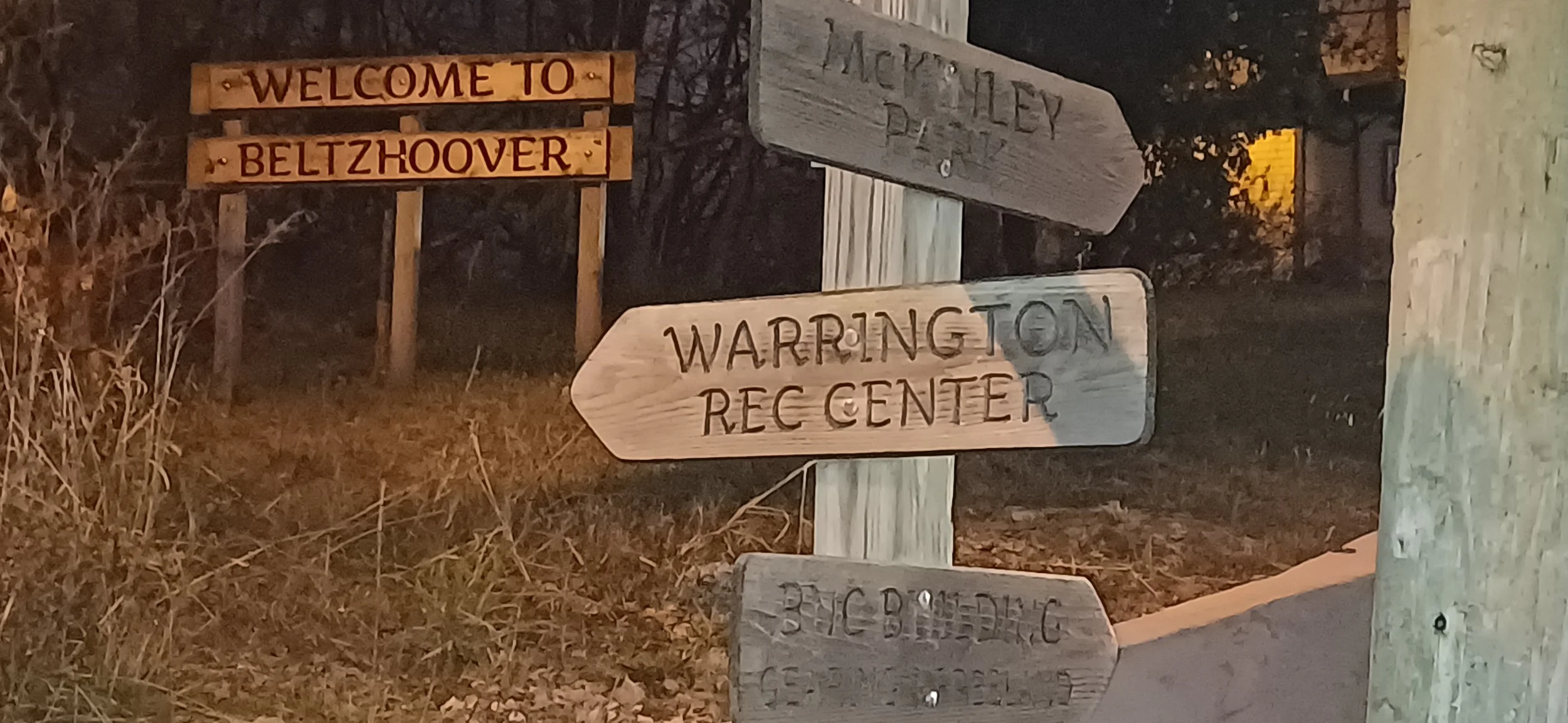 Wooden signs at night, one reads 'Welcome to Beltzhoover', another indicates directions to Waring ton Recreation Center, and a partially visible sign below.