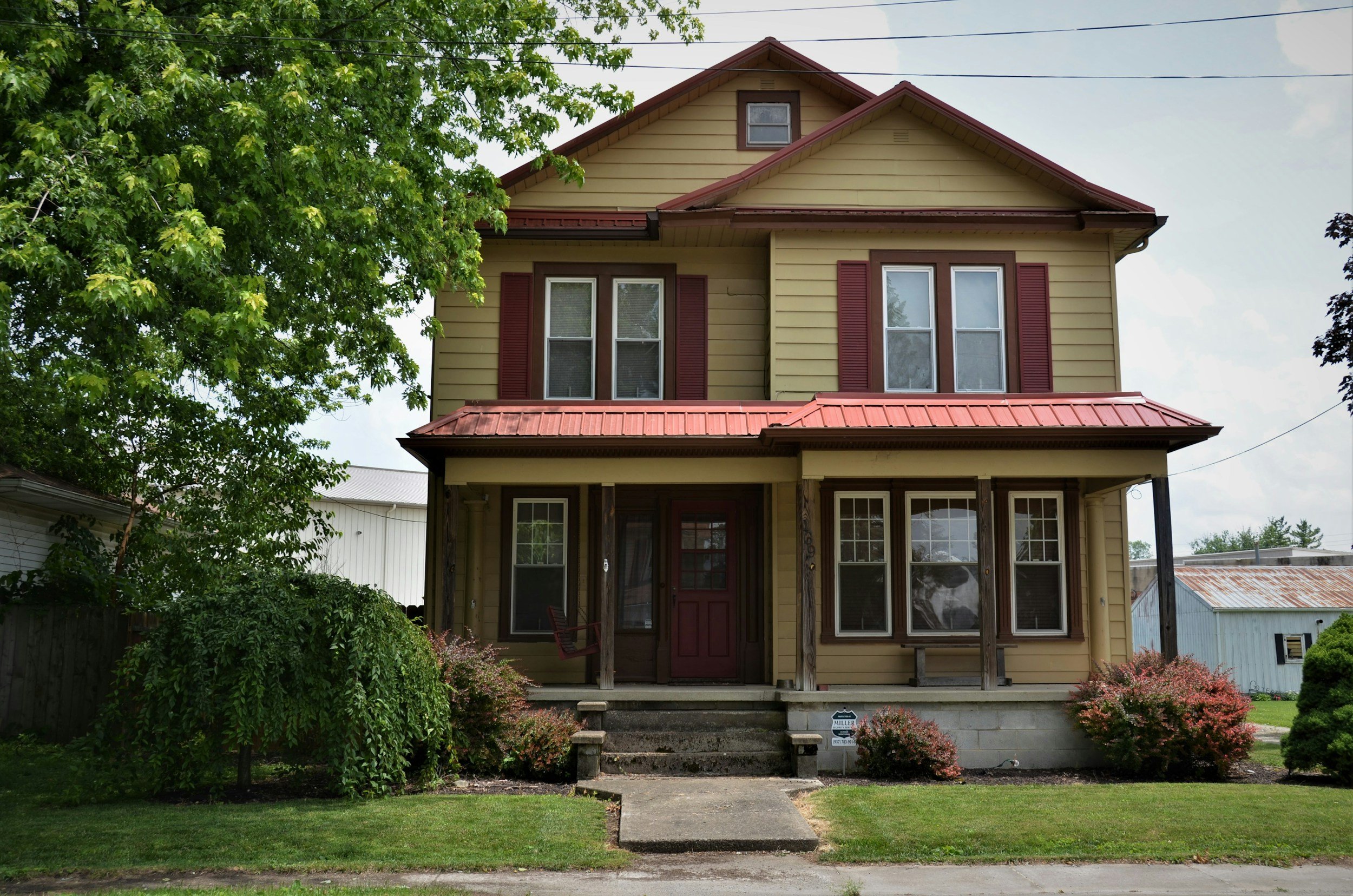 A two-story yellow house with red shutters and a red metal roof, front porch, and stairs, with a tree on the left and bushes in the front yard.