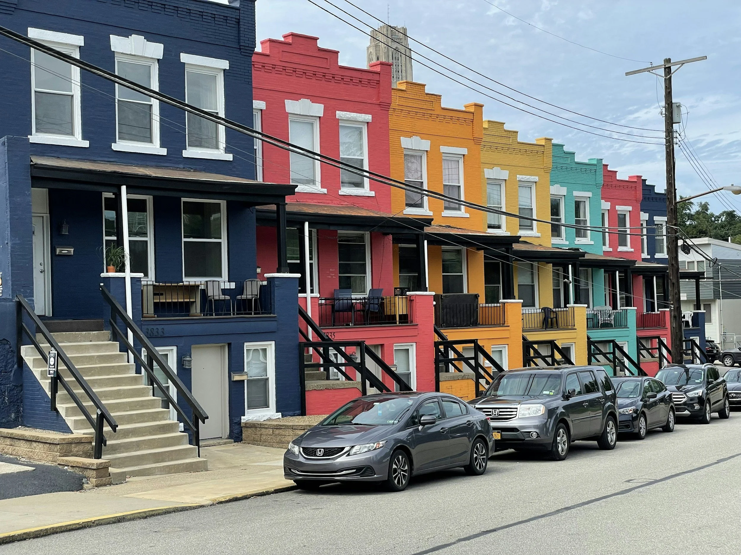 Colorful row of houses painted in blue, red, orange, yellow, teal, and dark blue with cars parked in front on a street, blue sky overhead.