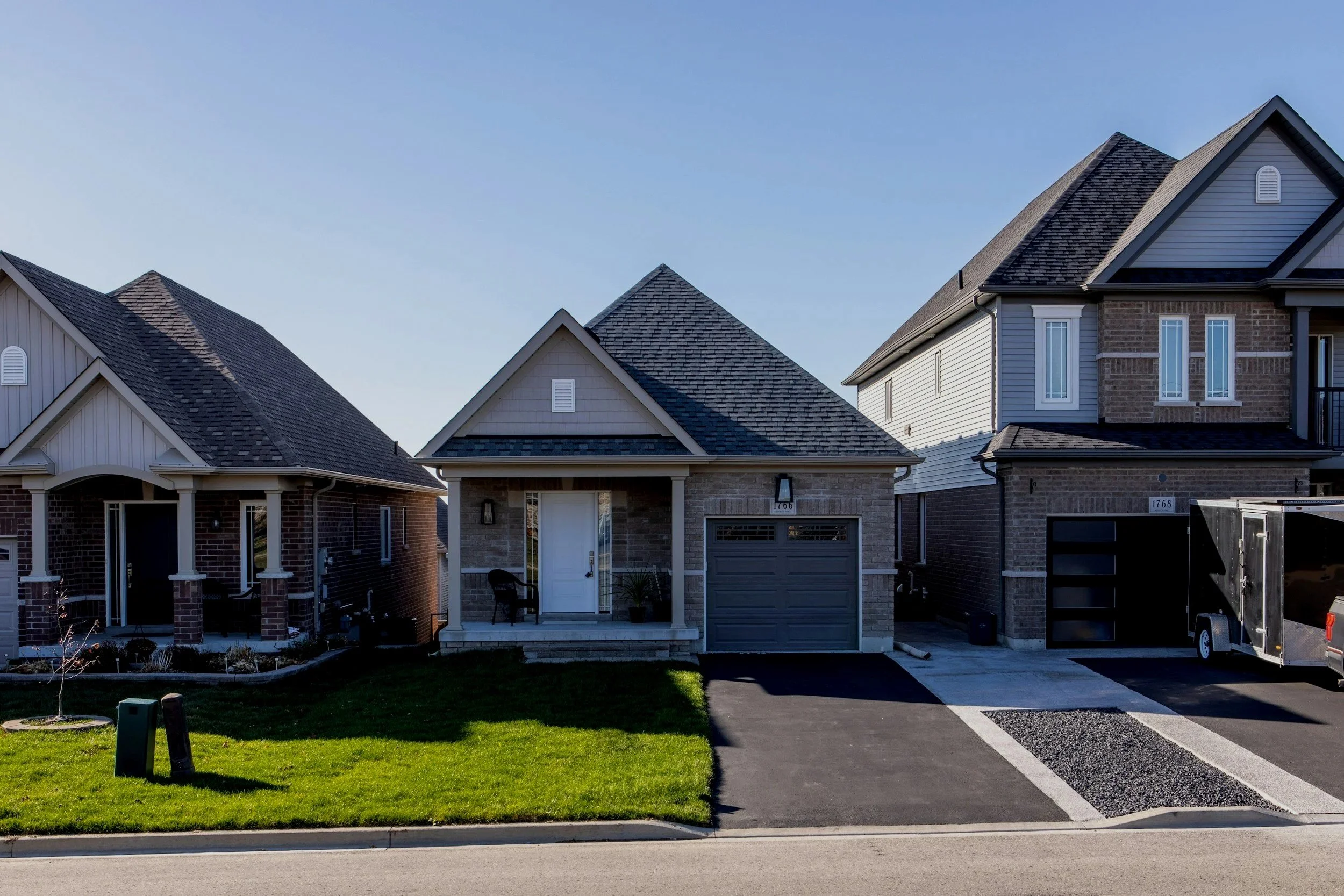 A row of three modern suburban houses with front yards, driveways, and garage doors, under a clear blue sky.