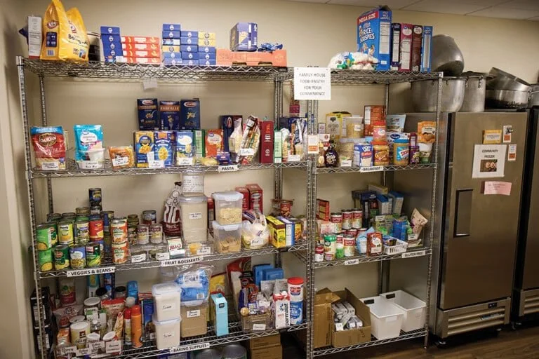 Metal shelves filled with various food and pantry items in a community kitchen or food pantry. Items include canned goods, boxes of pasta, cereal, baking supplies, and other non-perishable foods.