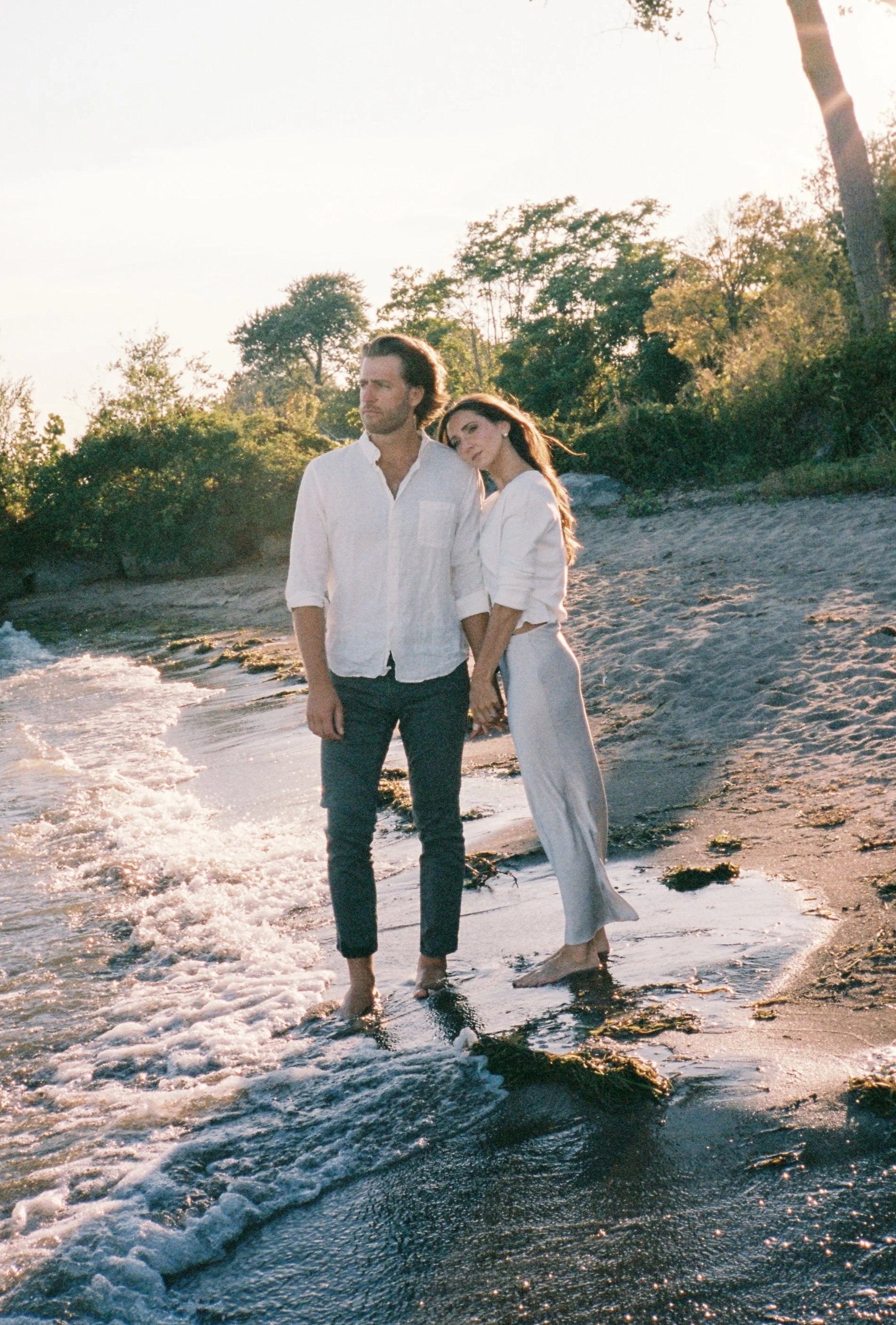 A man and woman holding hands on a beach, standing in shallow water, with trees and shrubs in the background, illuminated by warm sunlight.