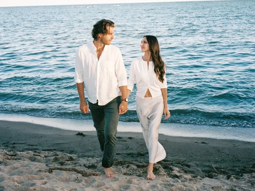 A man and woman walking barefoot on a beach near the ocean, holding hands and looking at each other.
