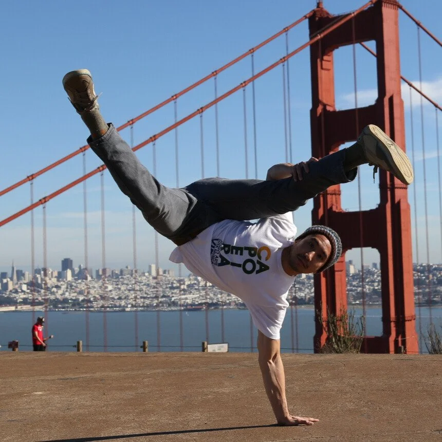 A man performing a one-handed handstand in front of the Golden Gate Bridge with a city skyline in the background on a clear day.