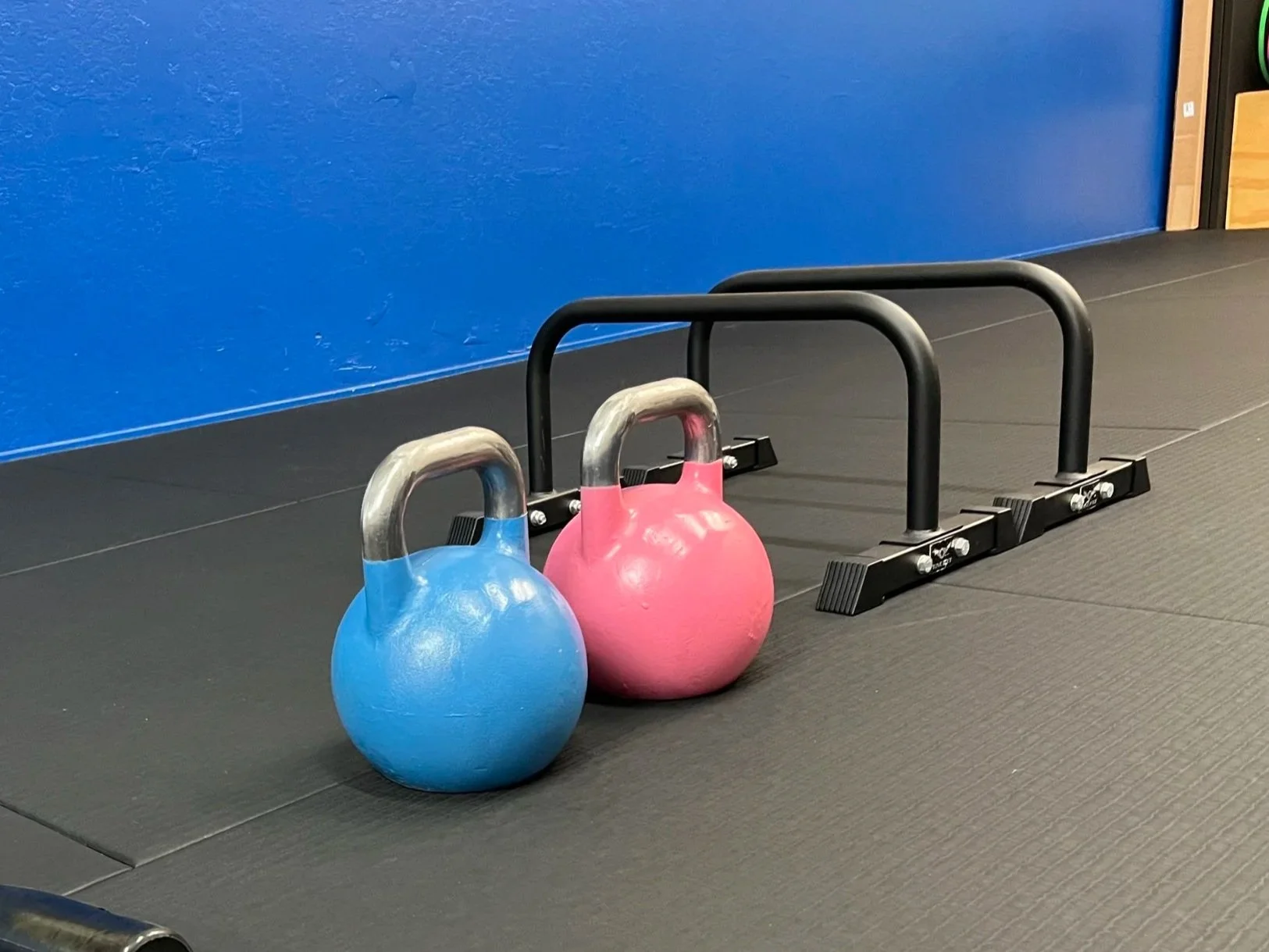Two kettlebells, one blue and one pink, placed on the floor next to a parallette push-up handle bar in a gym with black mats and a blue wall.