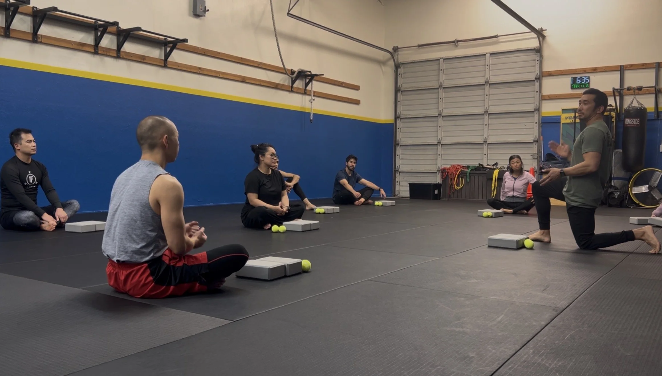 A group of six people sitting cross-legged on the floor in a gym, listening to a man in a green shirt who is kneeling and speaking to them. The gym has a black padded floor, a blue and yellow wall, and various equipment including a punching bag, wheels, and ropes.