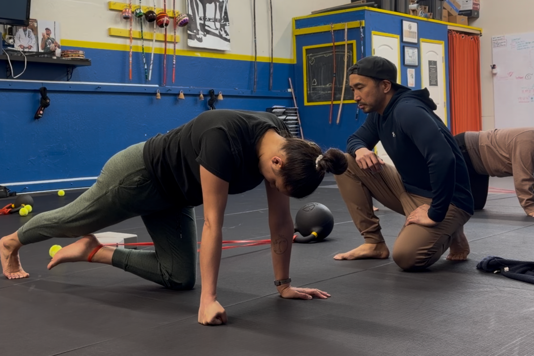 A woman practicing push-ups on a gym floor while a man kneels next to her instructing, with gym equipment of medicine balls, tennis balls, and ropes in the background.
