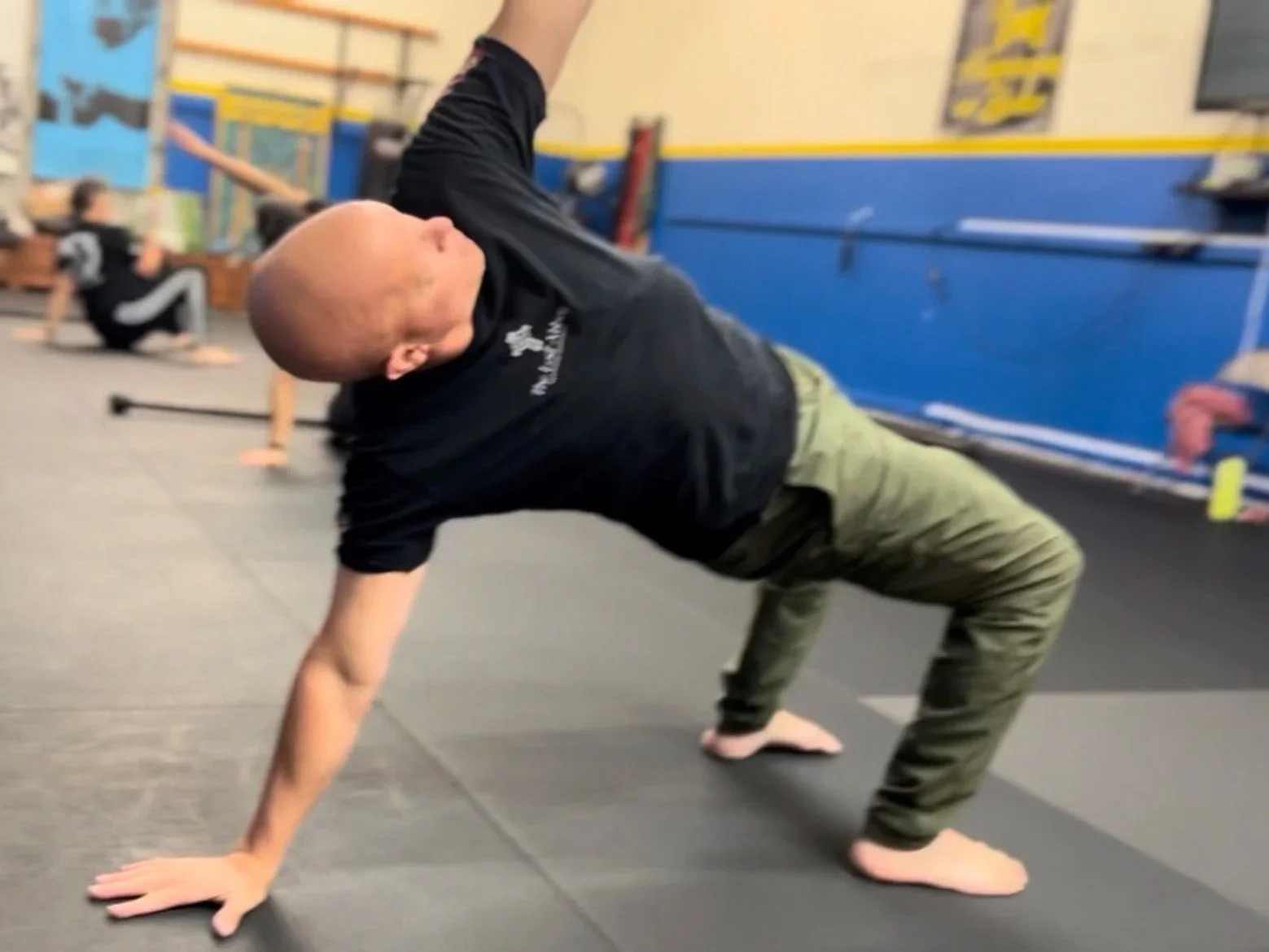 A man performing a yoga pose in a gym, balancing on one hand and one foot with the other arm extended upward, with other people doing similar exercises in the background.