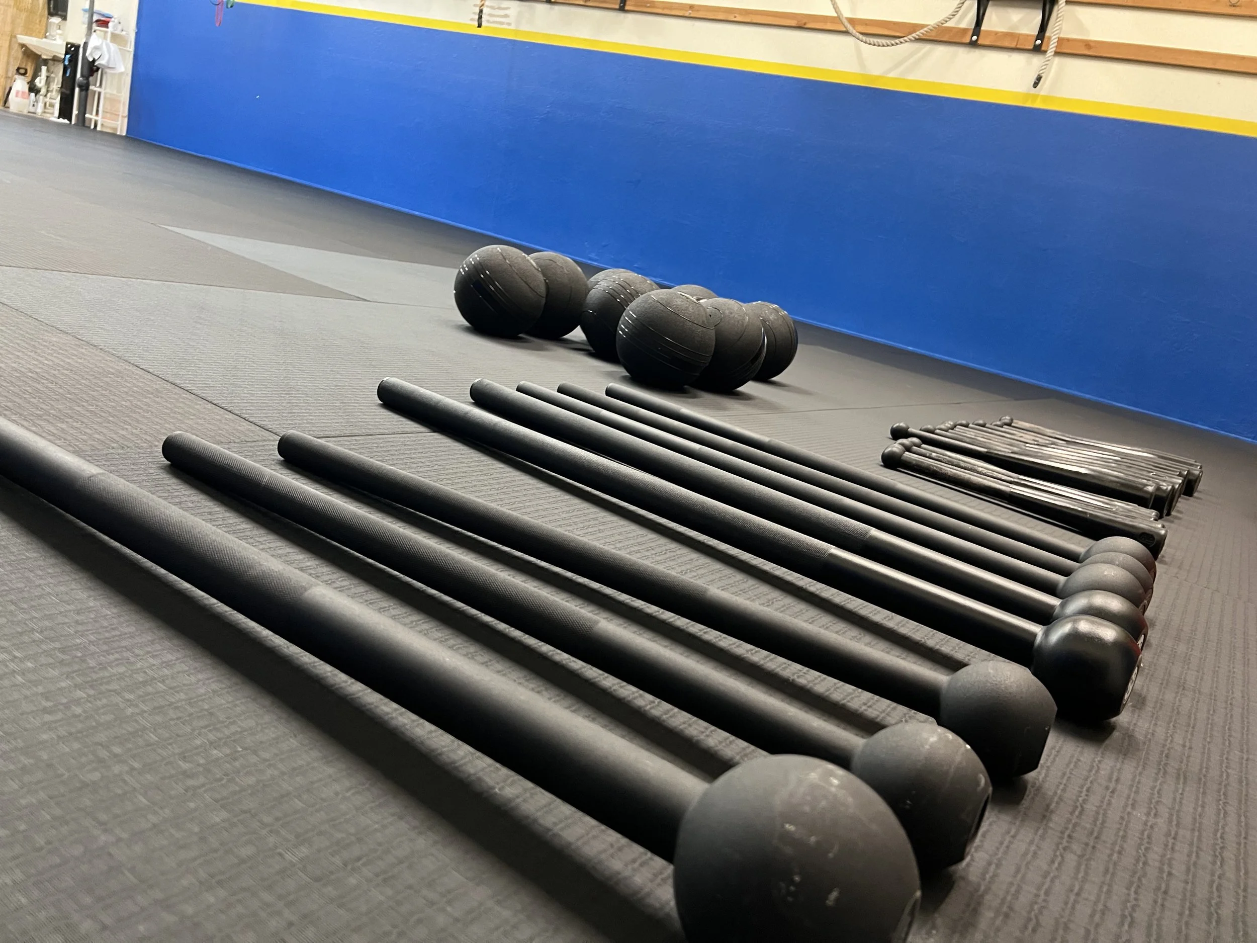 Flat lay of fitness equipment including black medicine balls, black battle ropes, and black push-up bars on a gym floor, with a blue wall in the background.