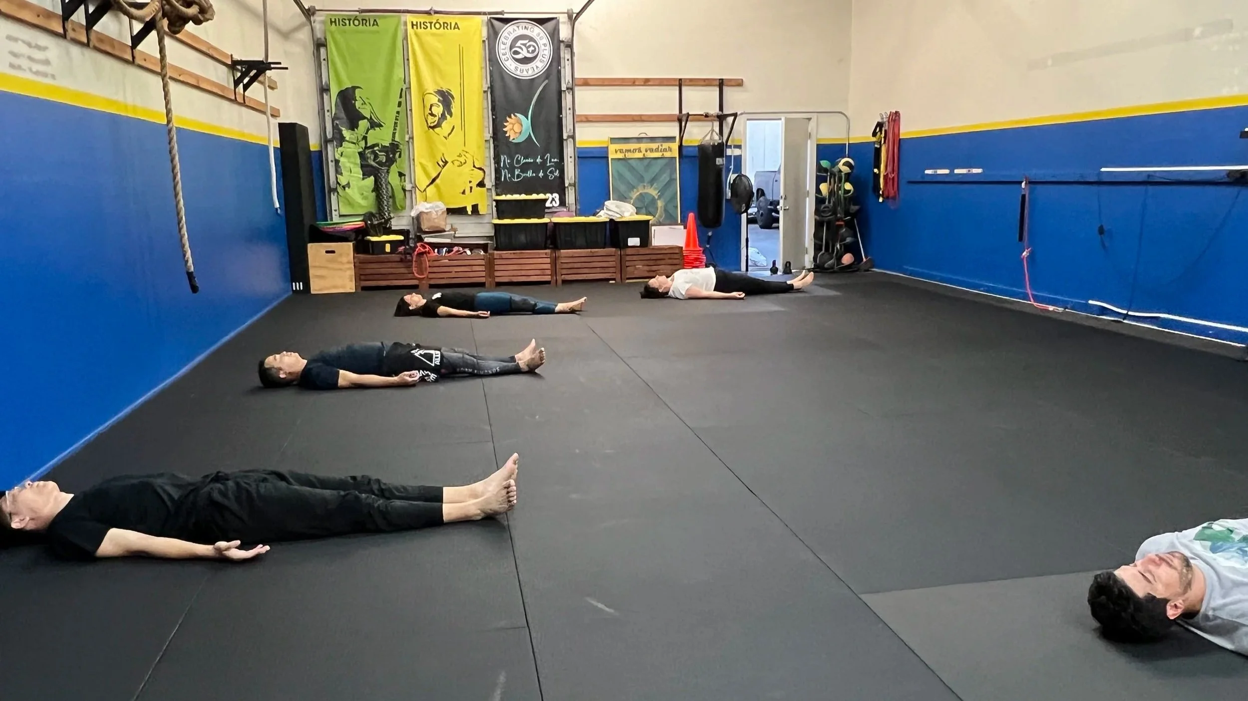 People lying on the floor in a gym or martial arts training room with black mats, colorful banners on the wall, free weights, and fitness equipment in the background.